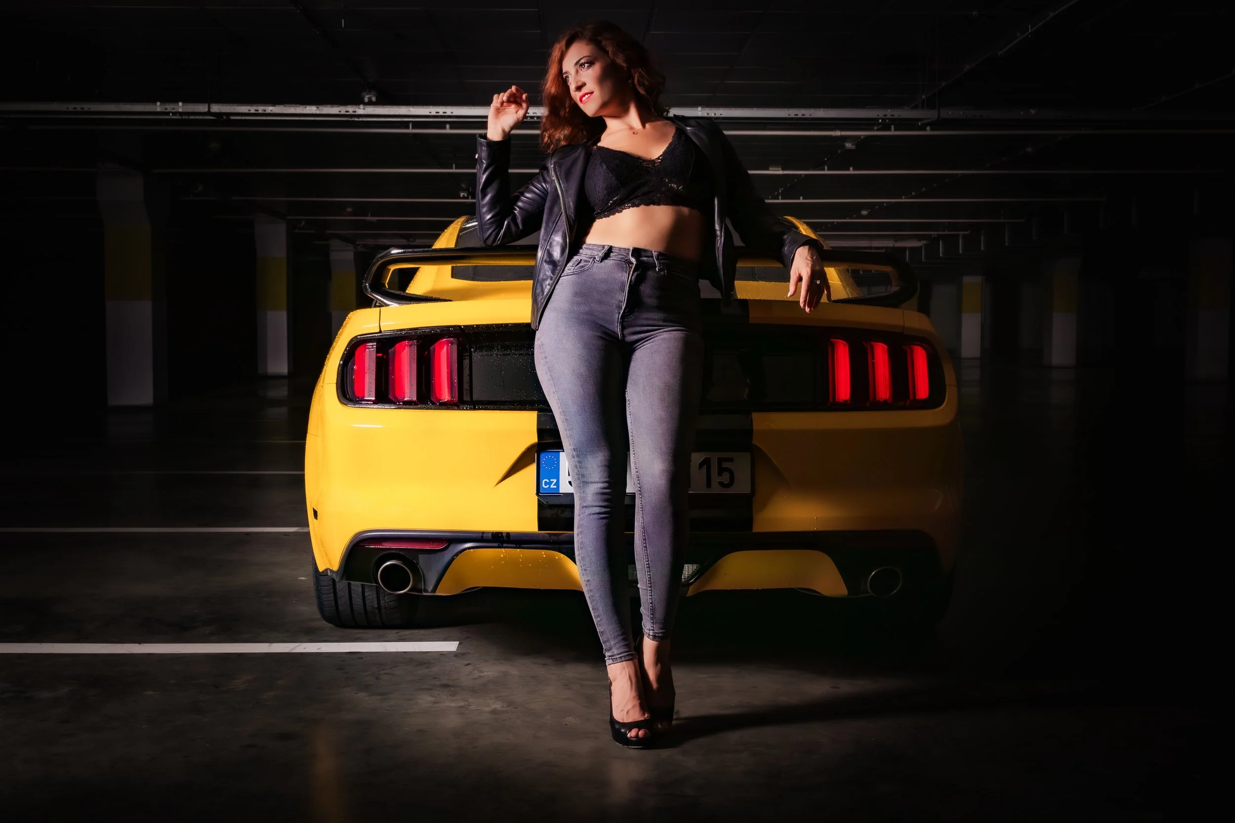 A woman in a black leather jacket, black crop top, and gray jeans standing in front of a yellow sports car inside a parking garage.