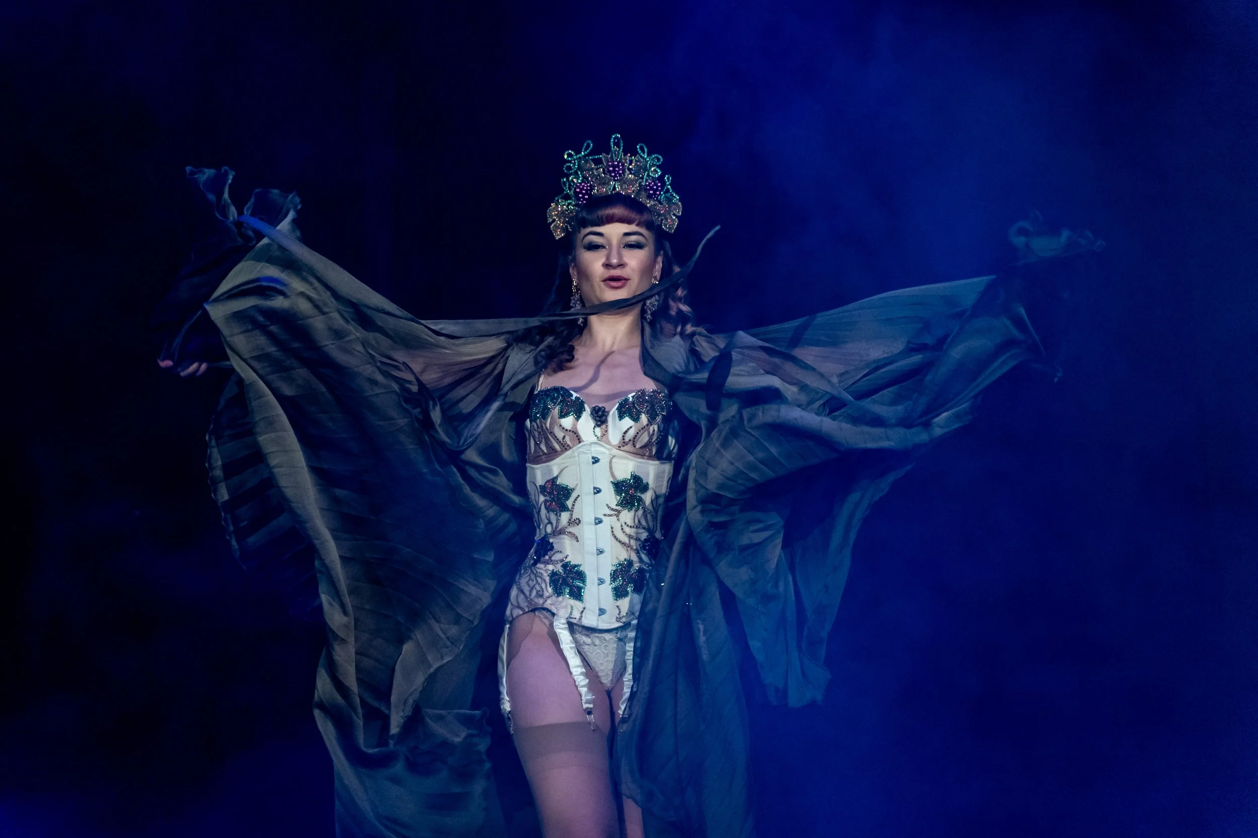 A woman wearing an ornate crown and elaborate lingerie holds large flowing black fabric in a dark setting with blue lighting.