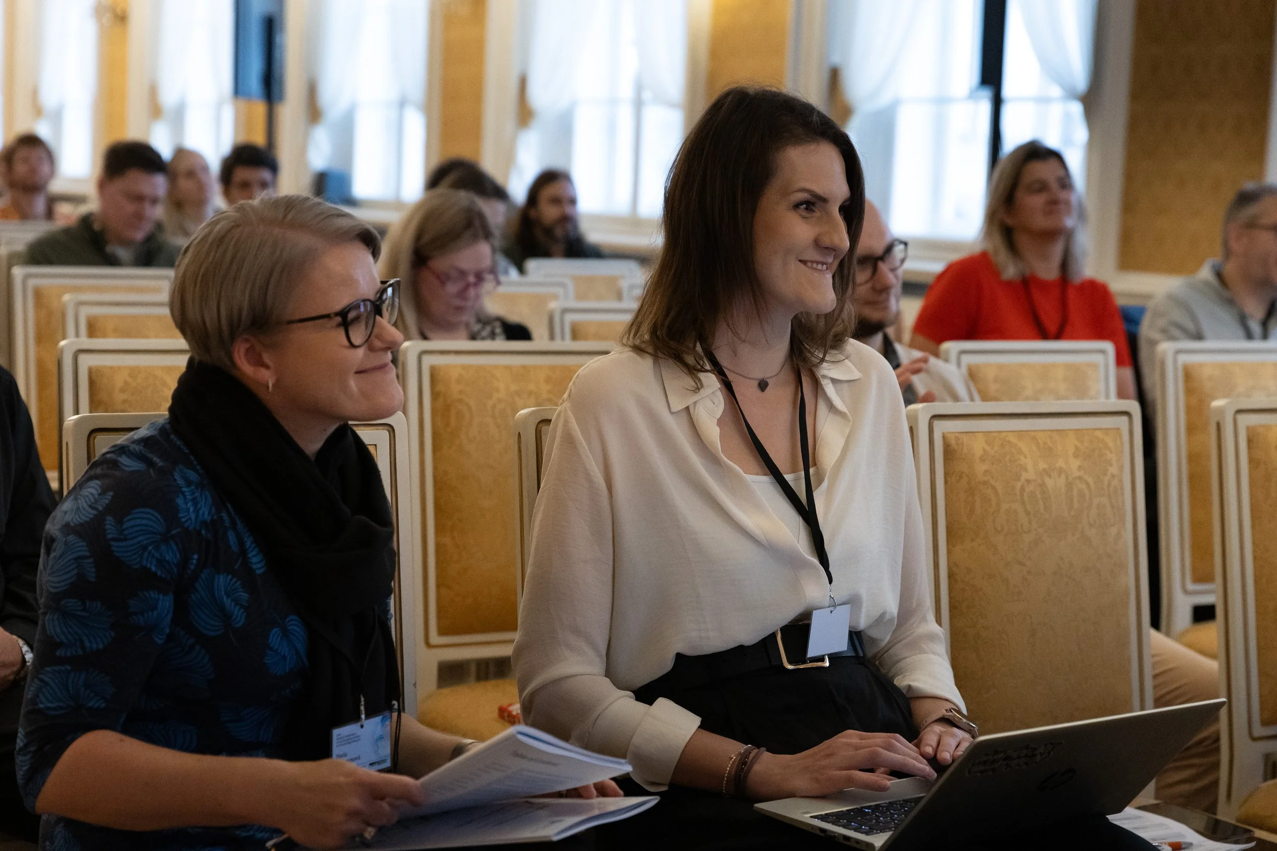 Two women sitting in an audience at a conference, smiling, with one woman using a laptop and the other holding papers.