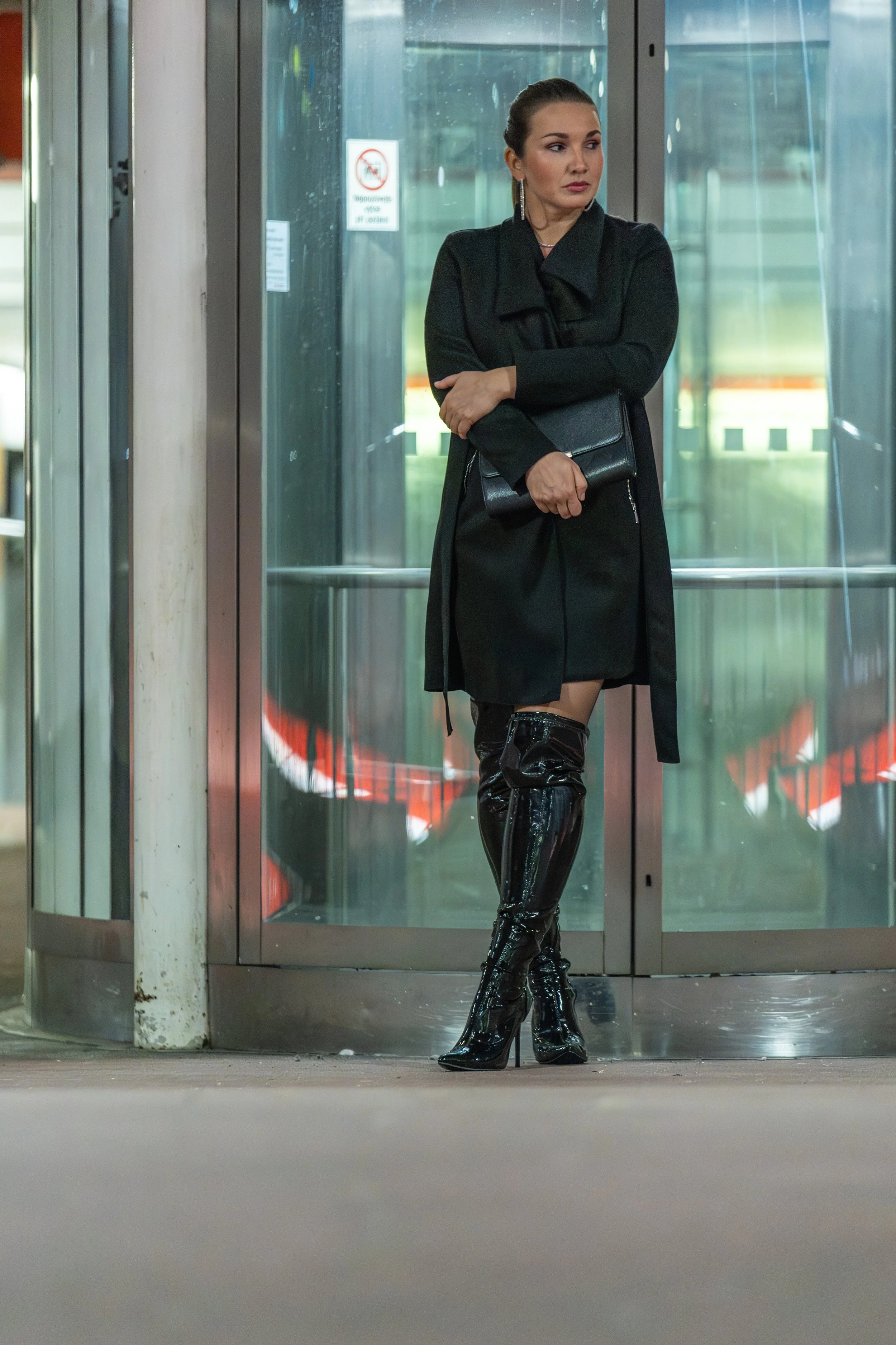 A woman standing outside an elevator wearing a black coat, shiny black thigh-high boots, and holding a black clutch.