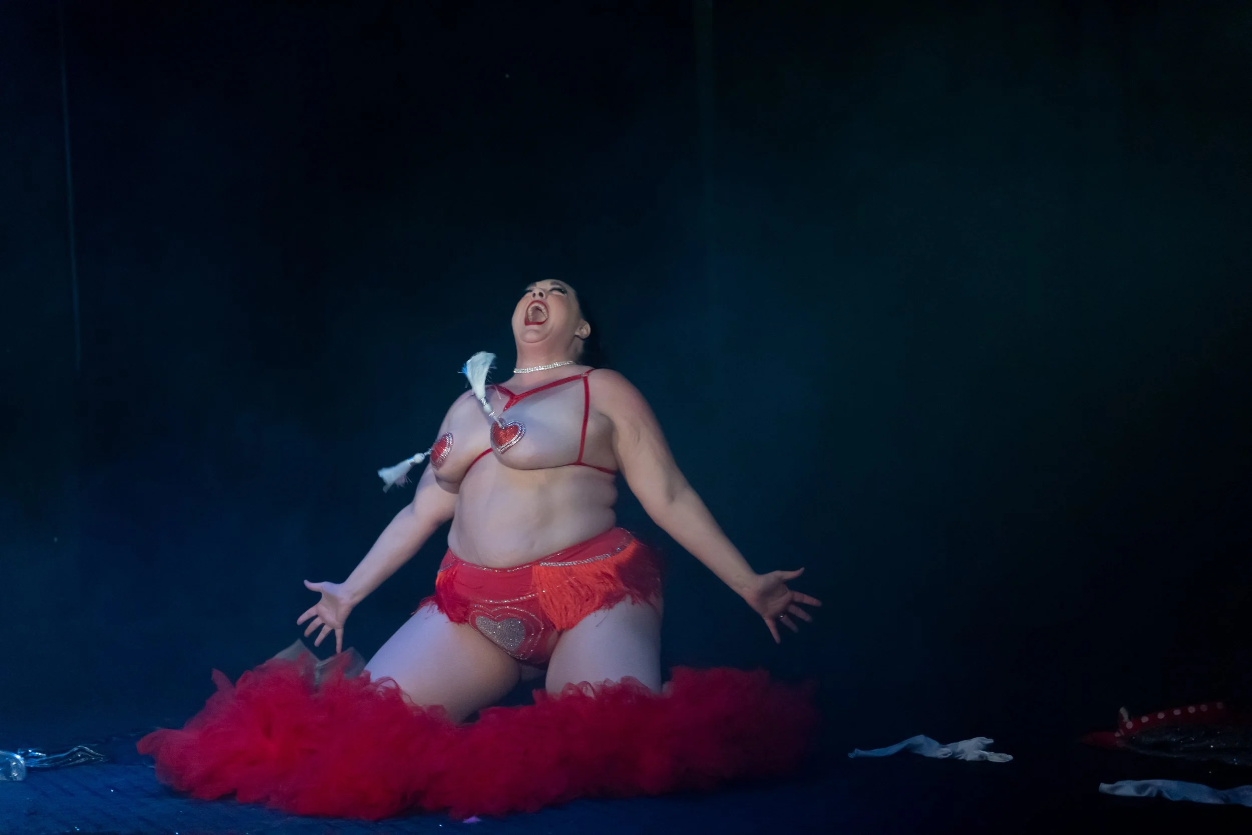 A woman dressed in a revealing costume with red feathers, heart-shaped pasties, and a feathered headpiece, kneeling with her arms outstretched and mouth open in a dramatic pose on a dark stage.