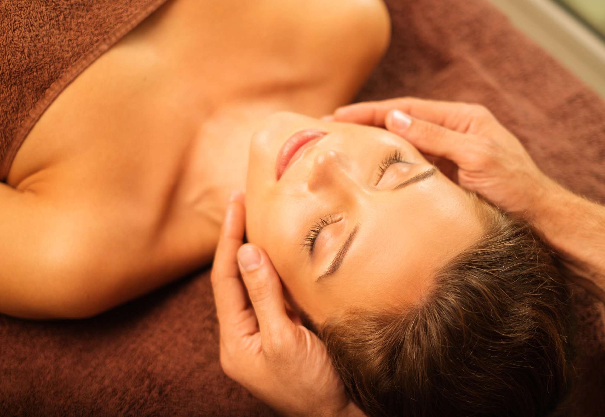 A woman is lying down peacefully with her eyes closed as a person gives her a head massage on a brown surface.