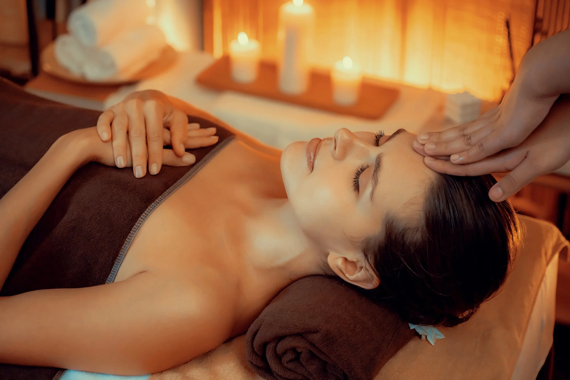 A woman lying on a massage table receiving a facial massage in a spa setting with warm lighting, candles, and towels.