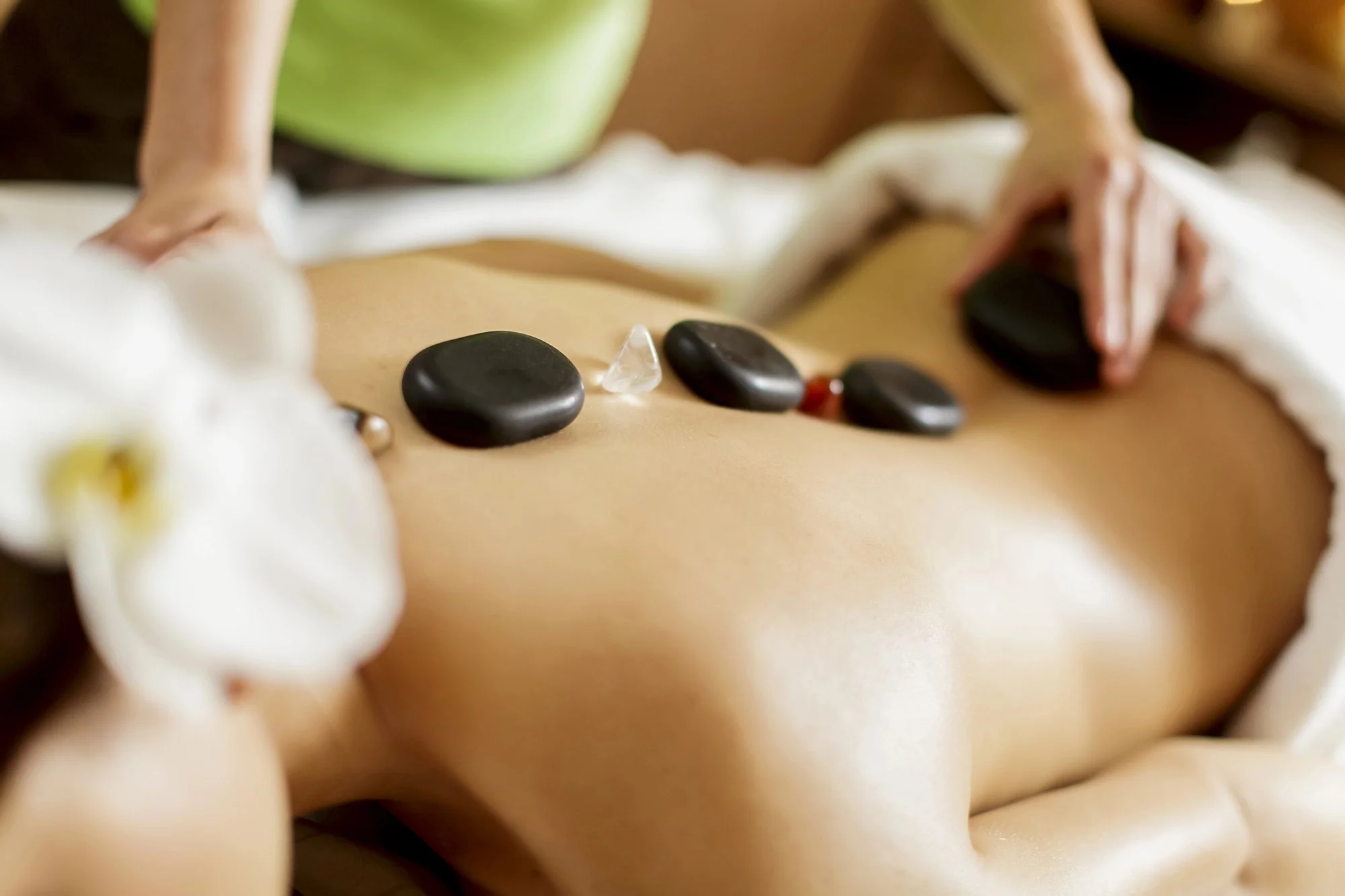 A person receiving a hot stone massage, with smooth black stones placed on their back and a massage therapist's hands applying pressure.
