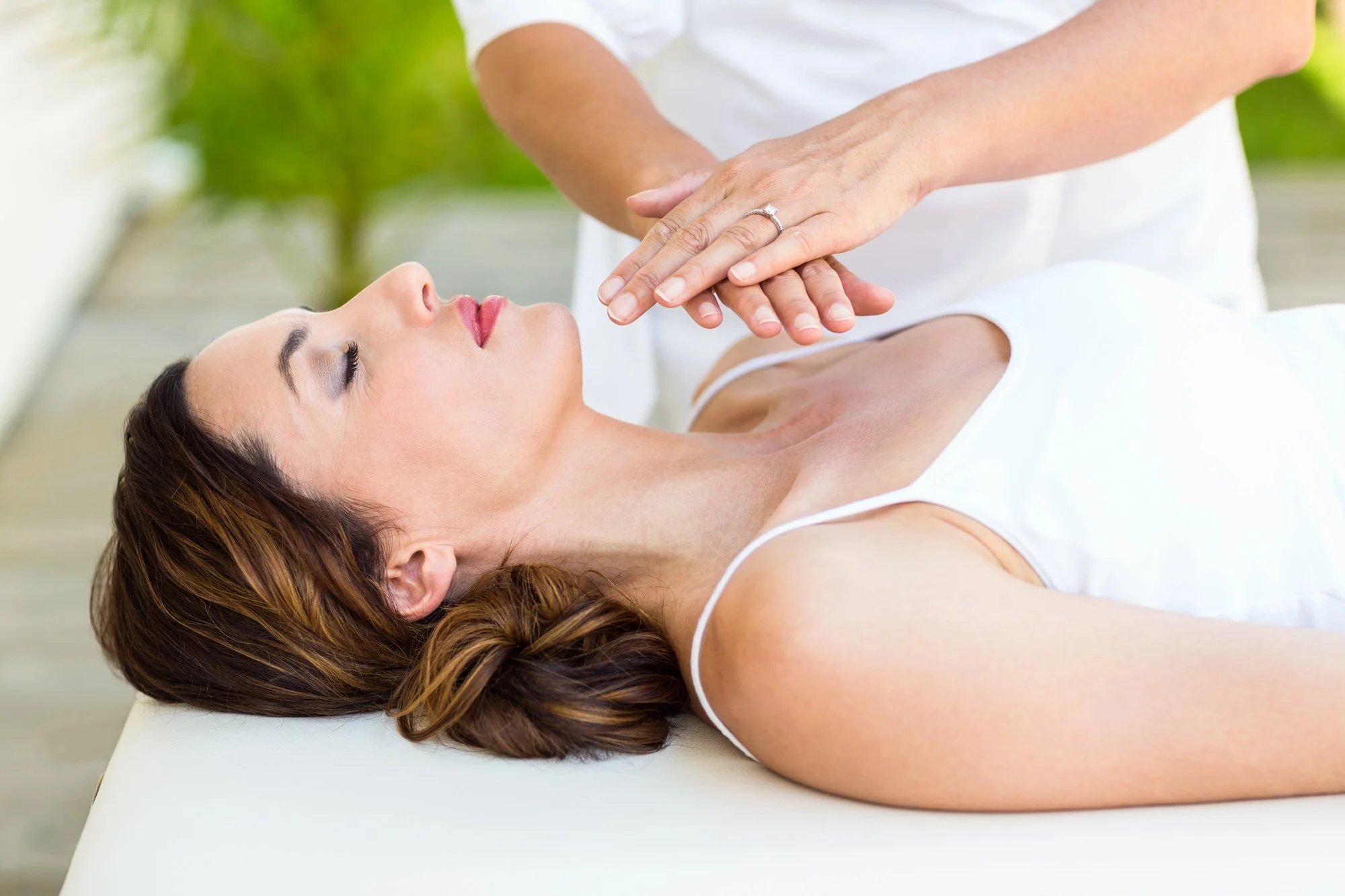 A woman receiving a Reiki session outdoors, lying on a treatment table with her eyes closed, as a practitioner places her hands above her chest.