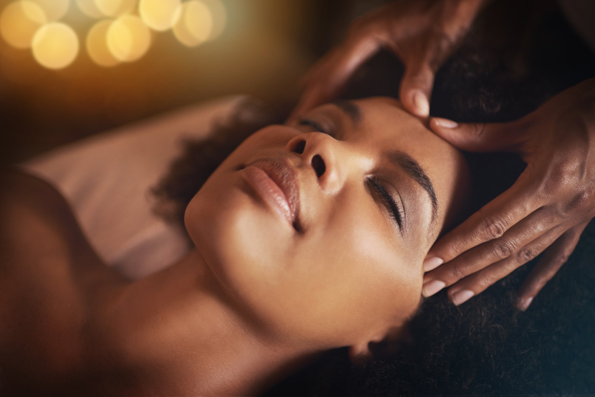 A woman receiving a massage, lying down with eyes closed, with a therapist's hands on her forehead, in a dimly lit room with warm lighting.