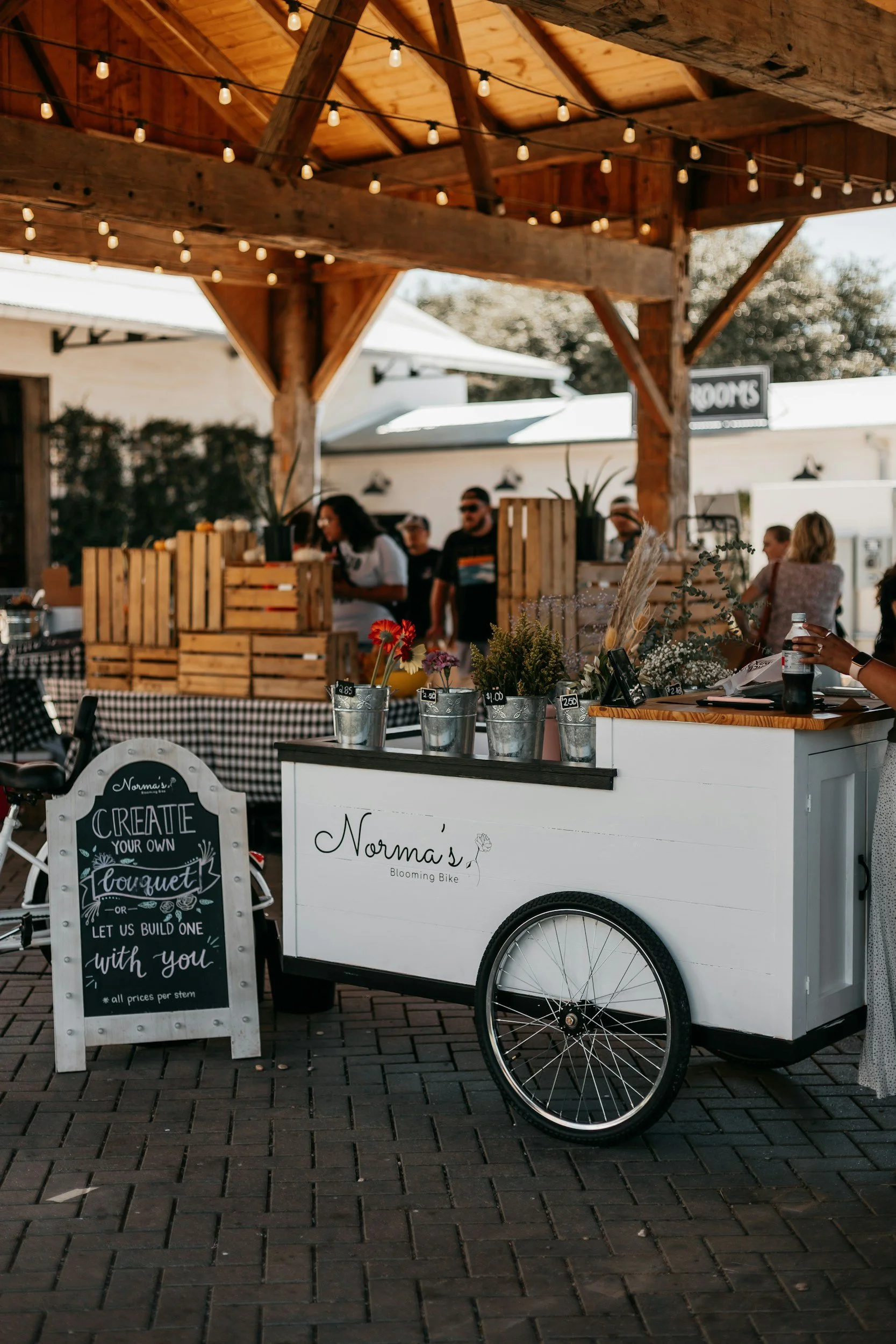 A white mobile flower cart labeled 'Norma's Blooming Bike' with potted flowers and a chalkboard sign in front promoting creating custom bouquets at a market or outdoor event.