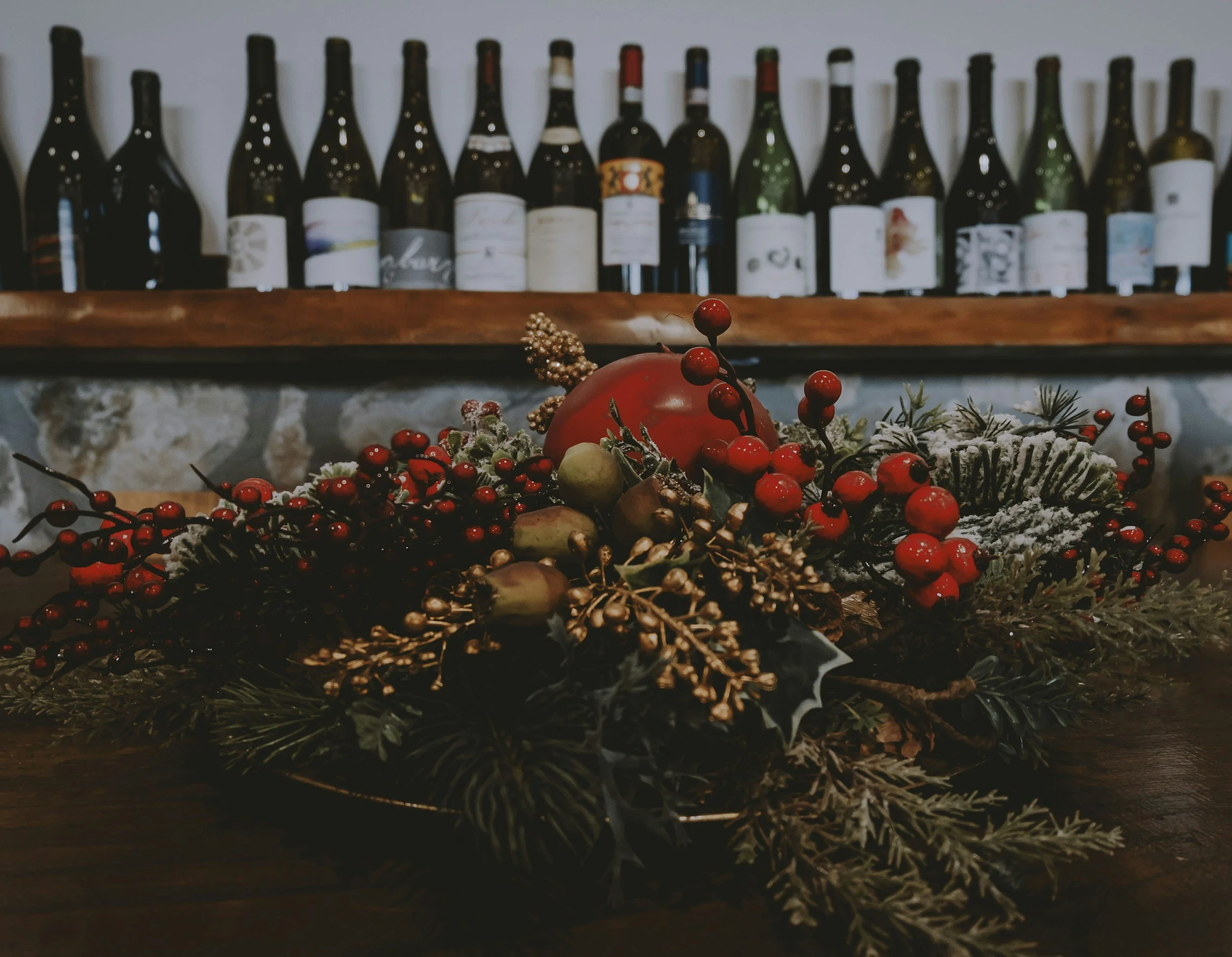 A holiday centerpiece with red berries, greenery, and gold accents resting on a wooden table, with a row of wine bottles on a shelf in the background.