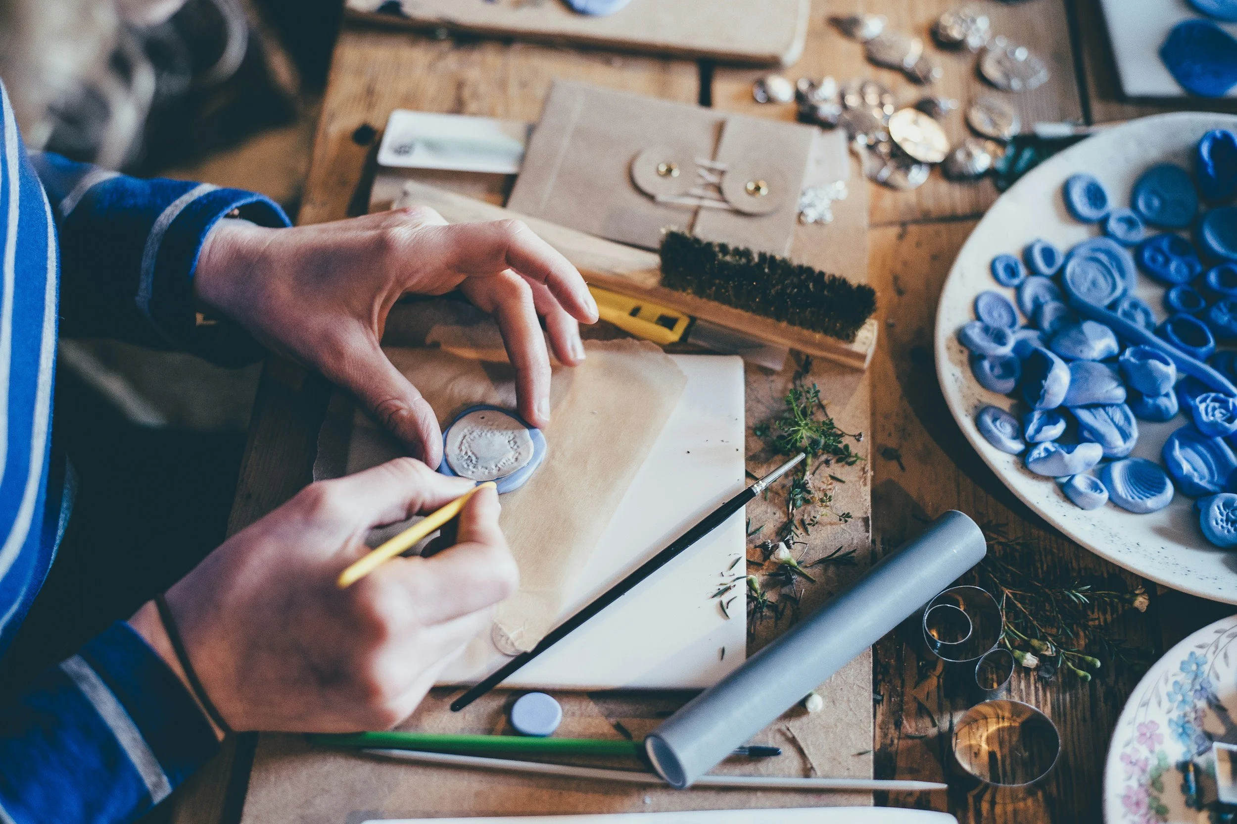A person working on a jewelry piece amidst various crafting supplies, including blue buttons, metallic rings, glue, and tools, on a wooden table.