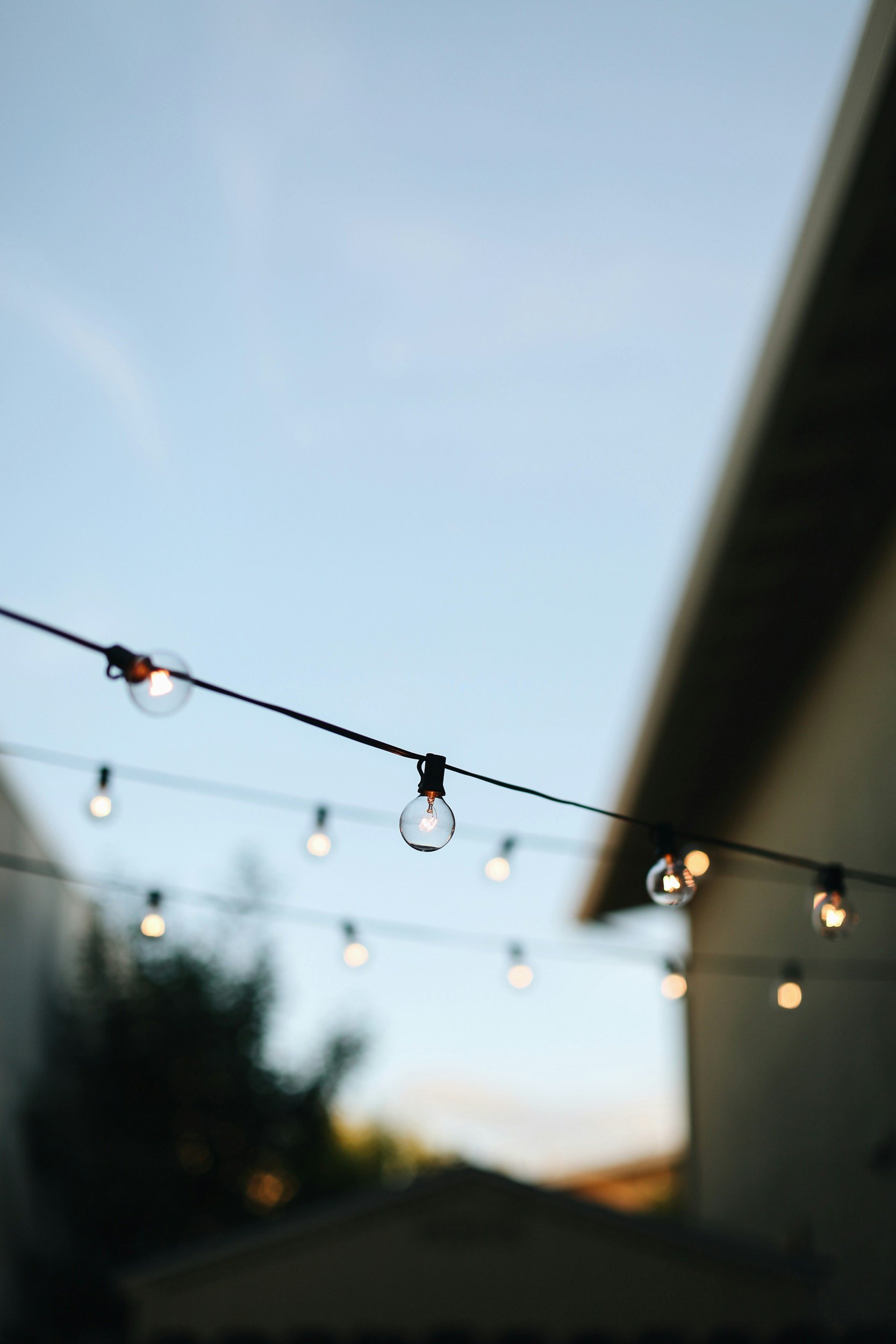 String lights hanging outdoors during twilight with a blurred house and trees in the background.