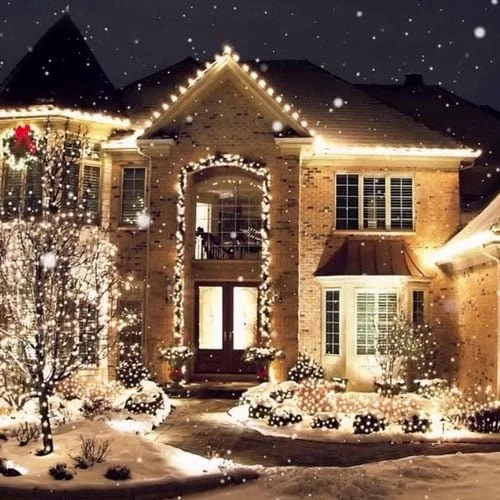 A large house decorated with Christmas lights and surrounded by snow, with snow falling in the evening.