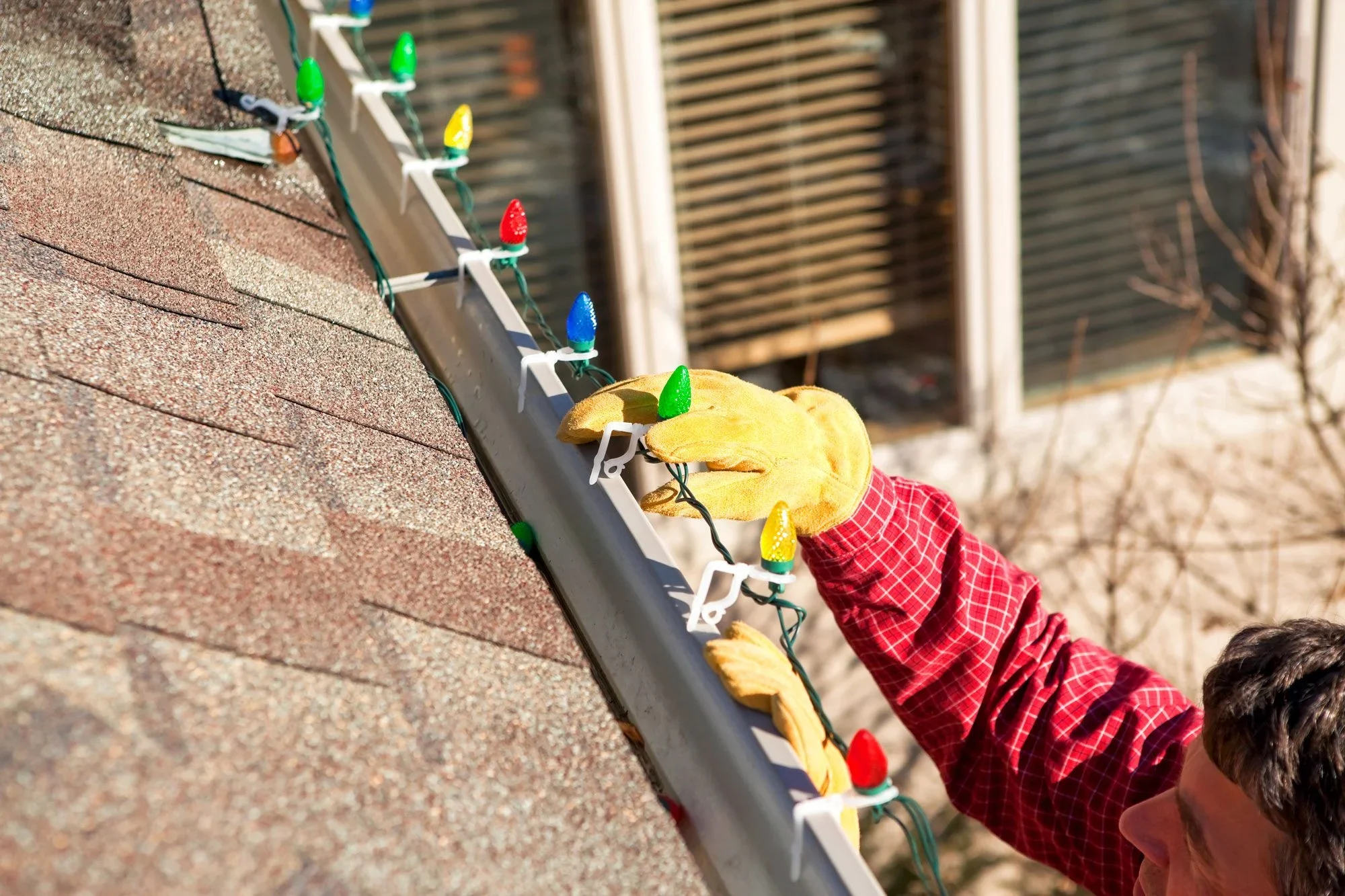 Person's hand, wearing a yellow glove and red plaid shirt, hanging colorful holiday lights on a house roof.