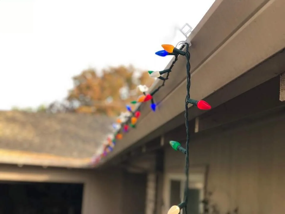 Close-up of colorful holiday string lights hanging from a roof edge against a blurred background of trees and sky.
