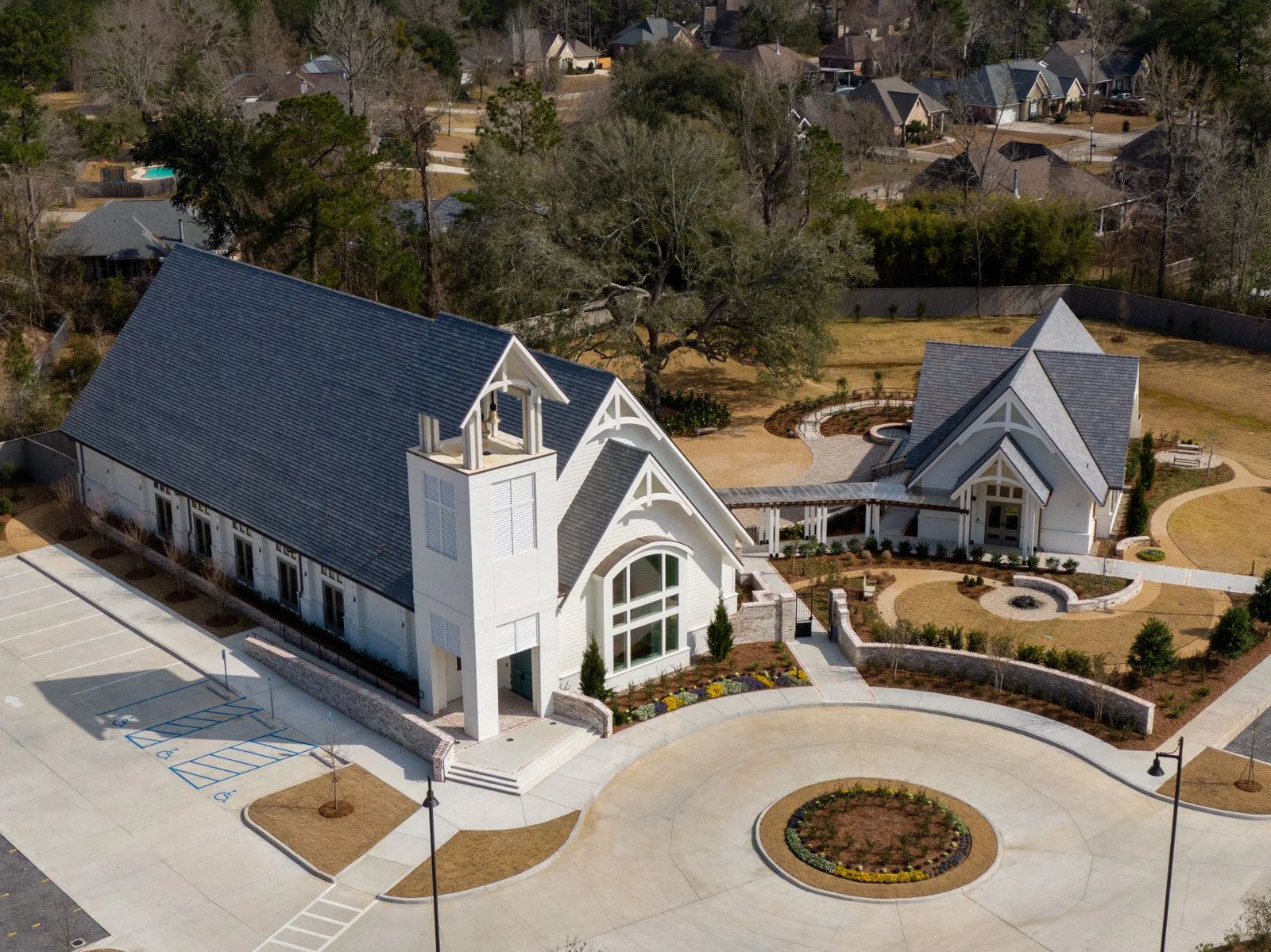 An aerial view of a church with a large parking lot, landscaped yard, and neighboring houses in the background.