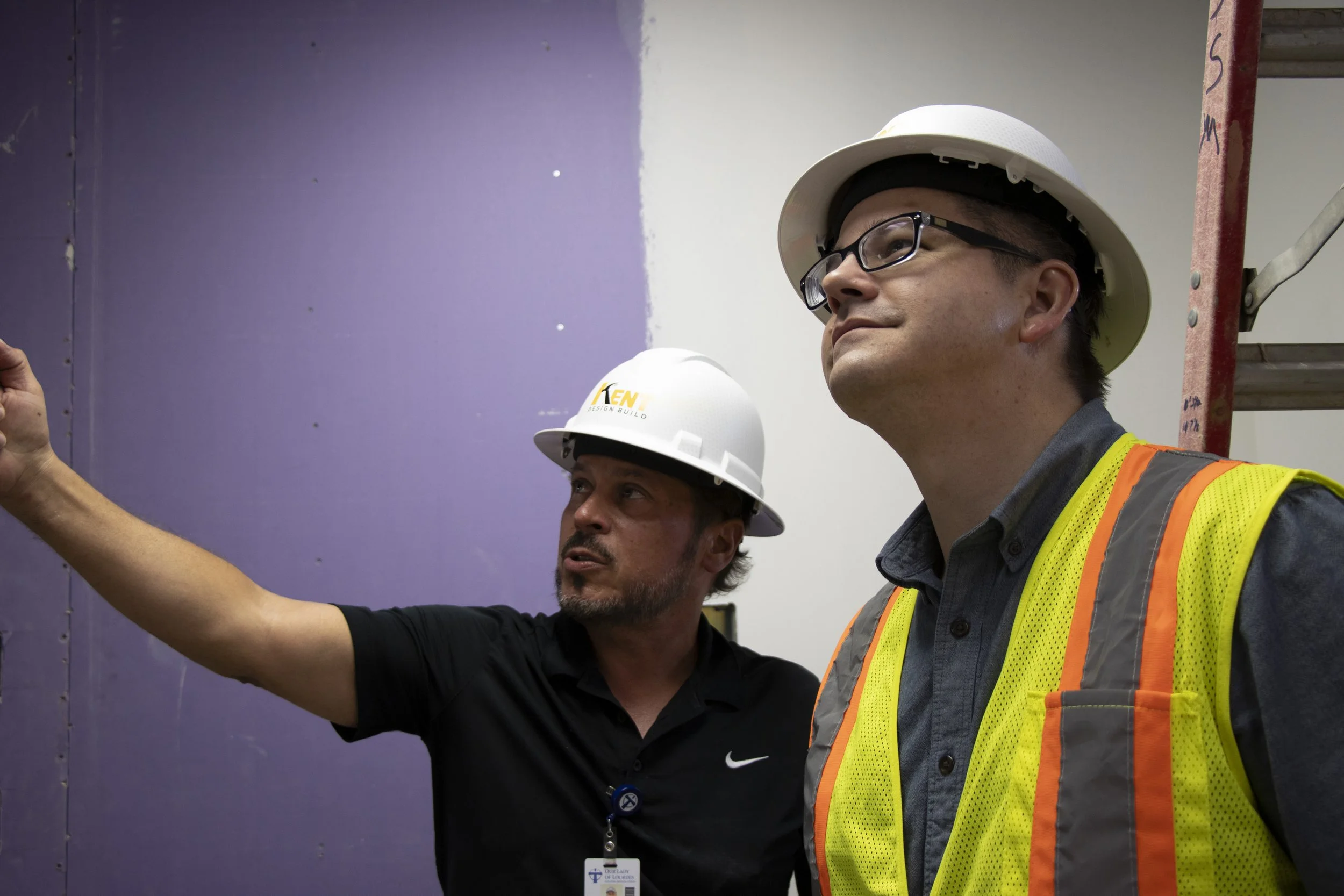 Two construction workers wearing hard hats and safety vests on a construction site, with one pointing at a wall.