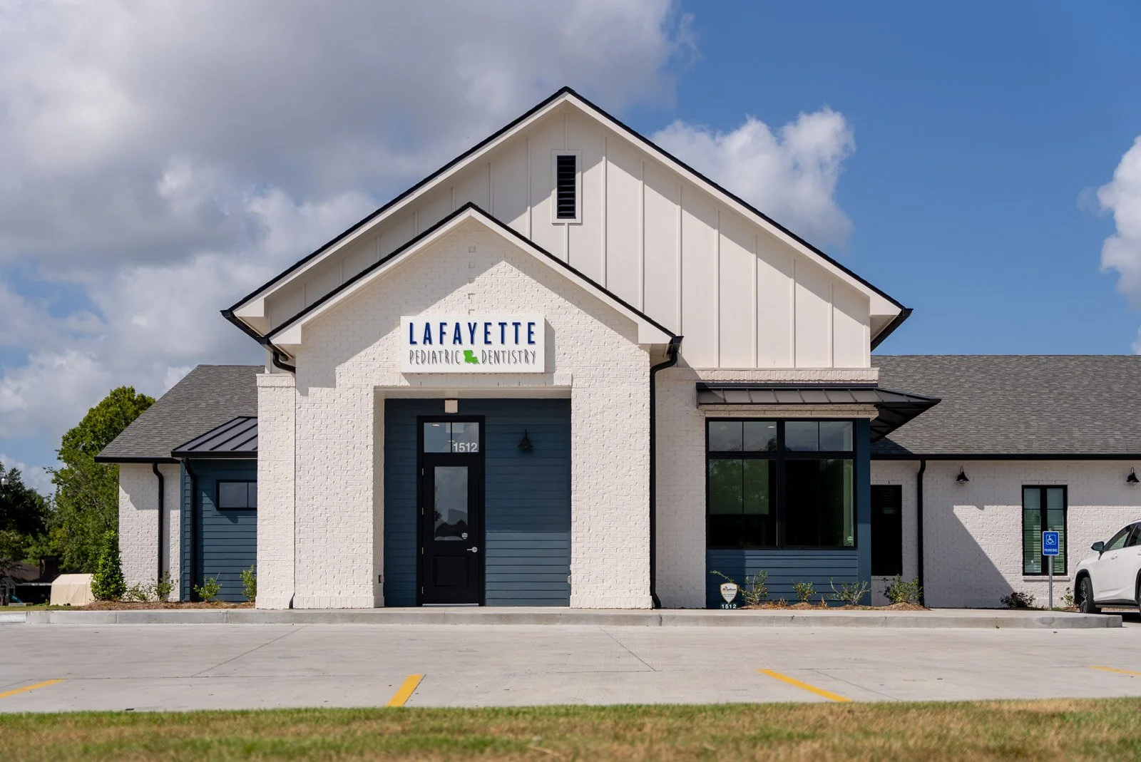 Exterior of Lafayette Pediatric Dentistry office with white brick walls, blue accents, and a sign reading Lafayette Pediatric Dentistry, parked cars, and blue sky with clouds.