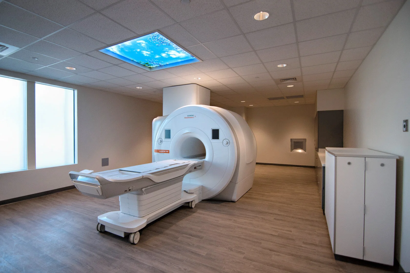 Empty hospital room with MRI machine, wood-like flooring, white cabinets, and windows with frosted glass.