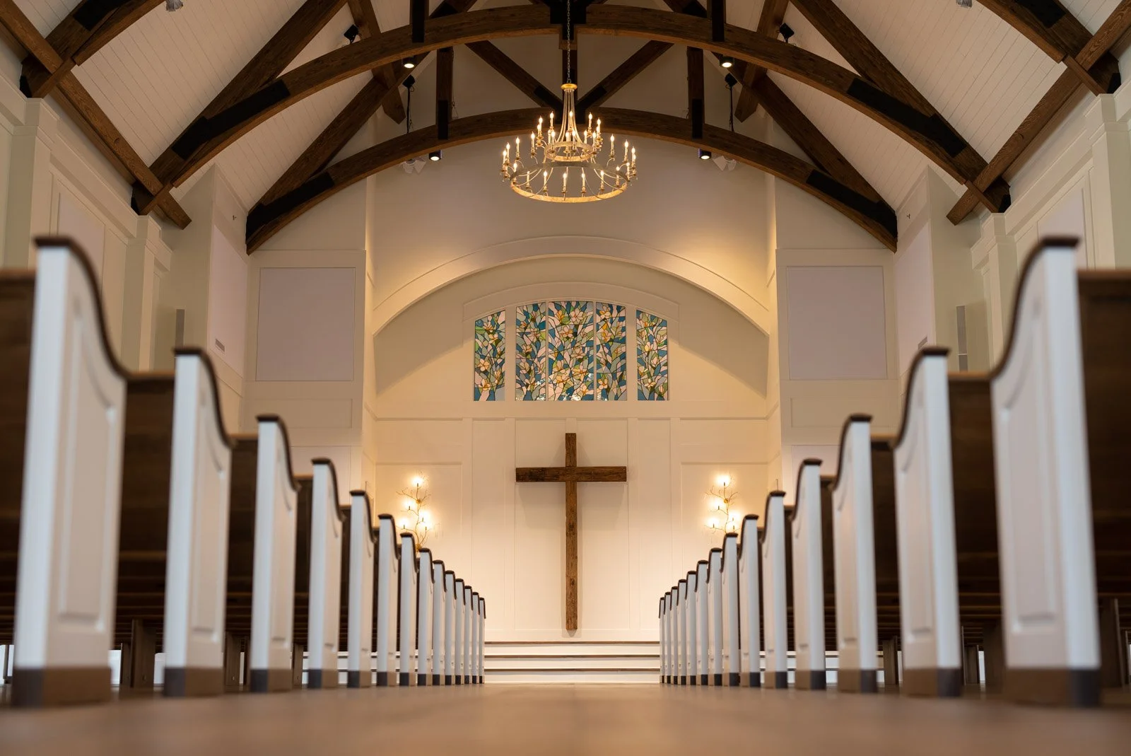 Interior of a church with white walls, wooden pews, a cross on the front wall, stained glass window above the altar, chandelier, and wall sconces.