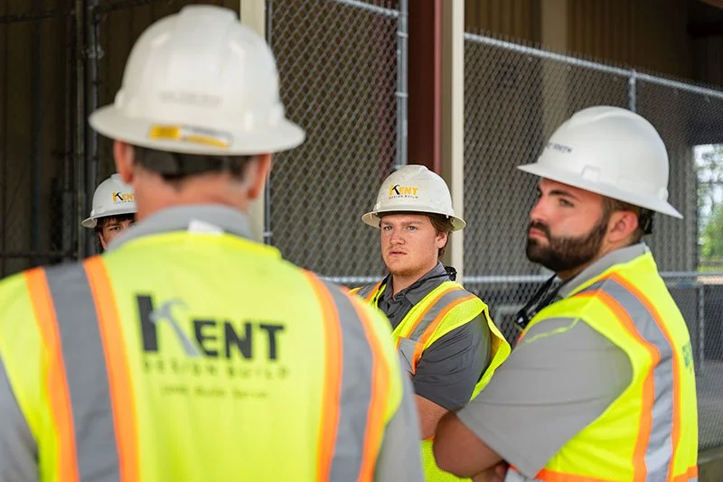 Group of construction workers wearing yellow reflective vests and white hard hats, standing outdoors near a construction site with a chain-link fence.