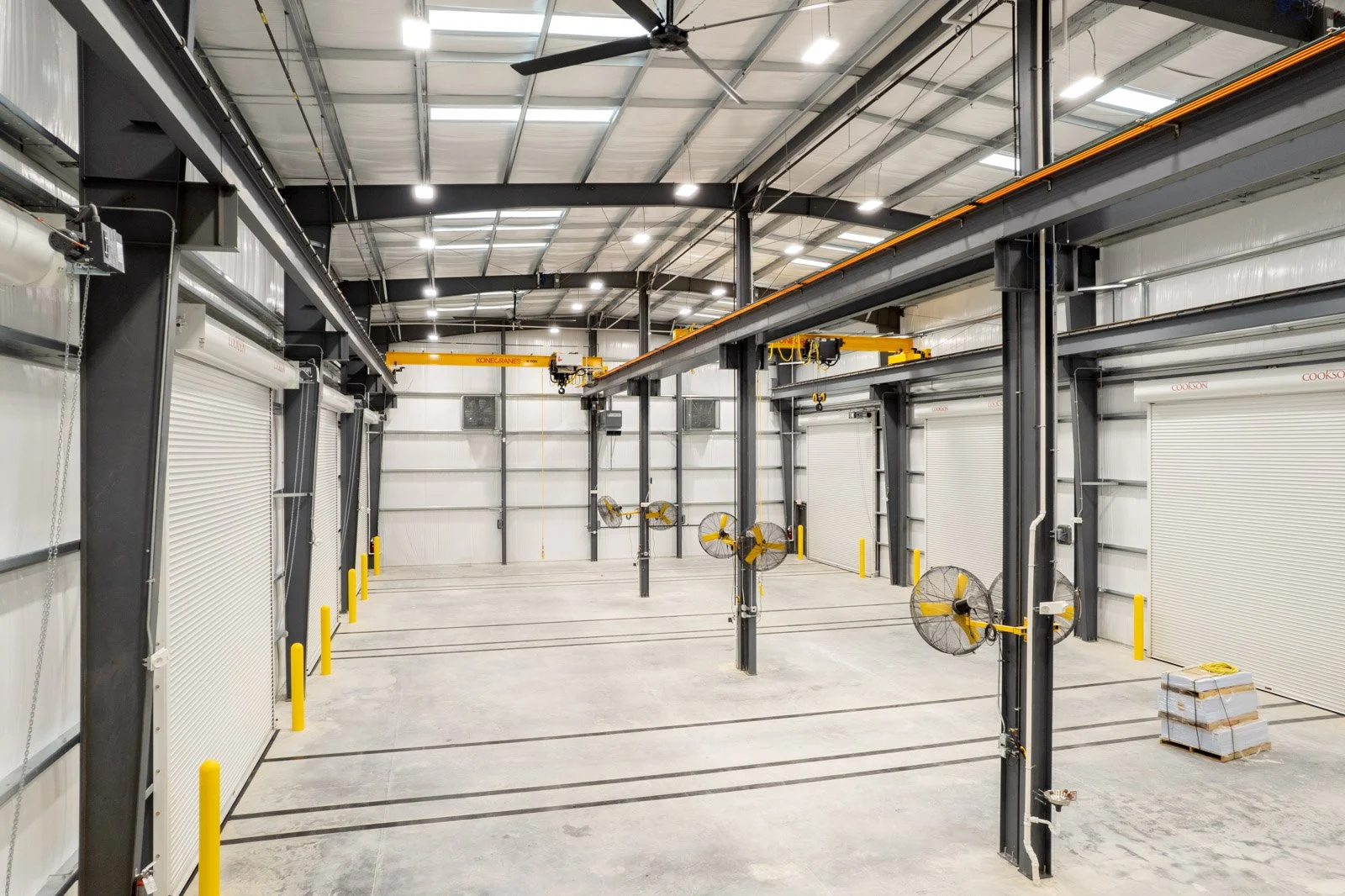 Empty industrial warehouse with large overhead doors, ceiling fans, and wall-mounted fans, with a pallet on floor and overhead cranes.