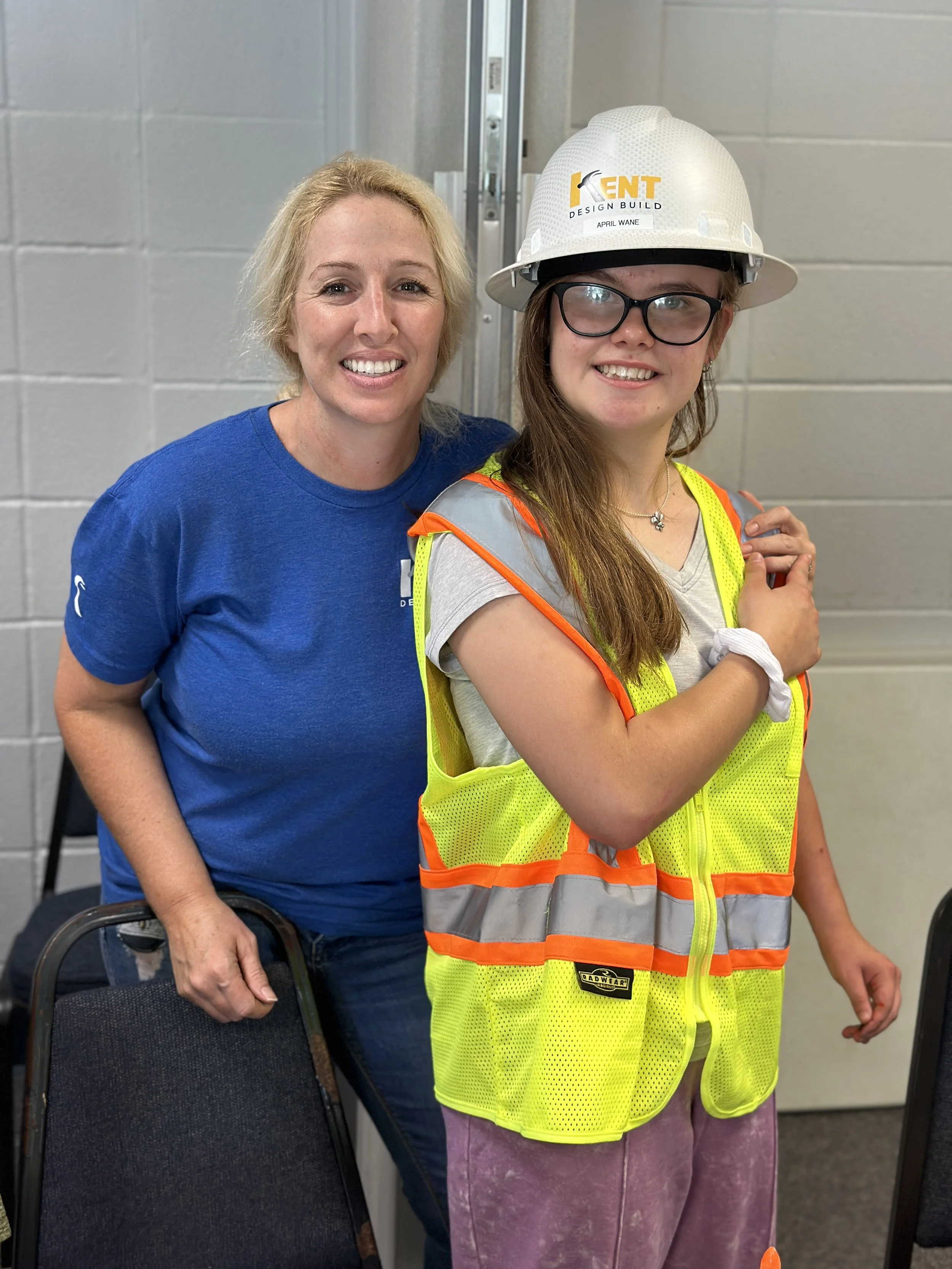 Two women standing indoor; one wears a blue shirt, the other a white safety helmet and a high-visibility vest, smiling.