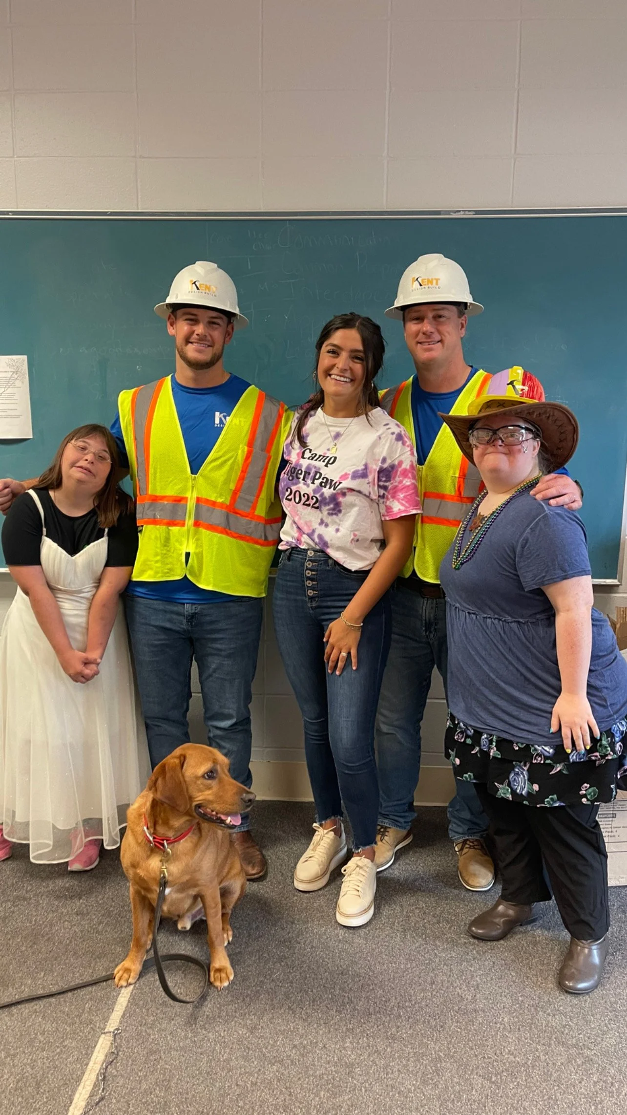 Group of five people, including three young adults, an older woman, and a young girl, standing with a dog in front of a chalkboard. The young adults are wearing construction helmets and safety vests. The girl is in a white dress, and the older woman 