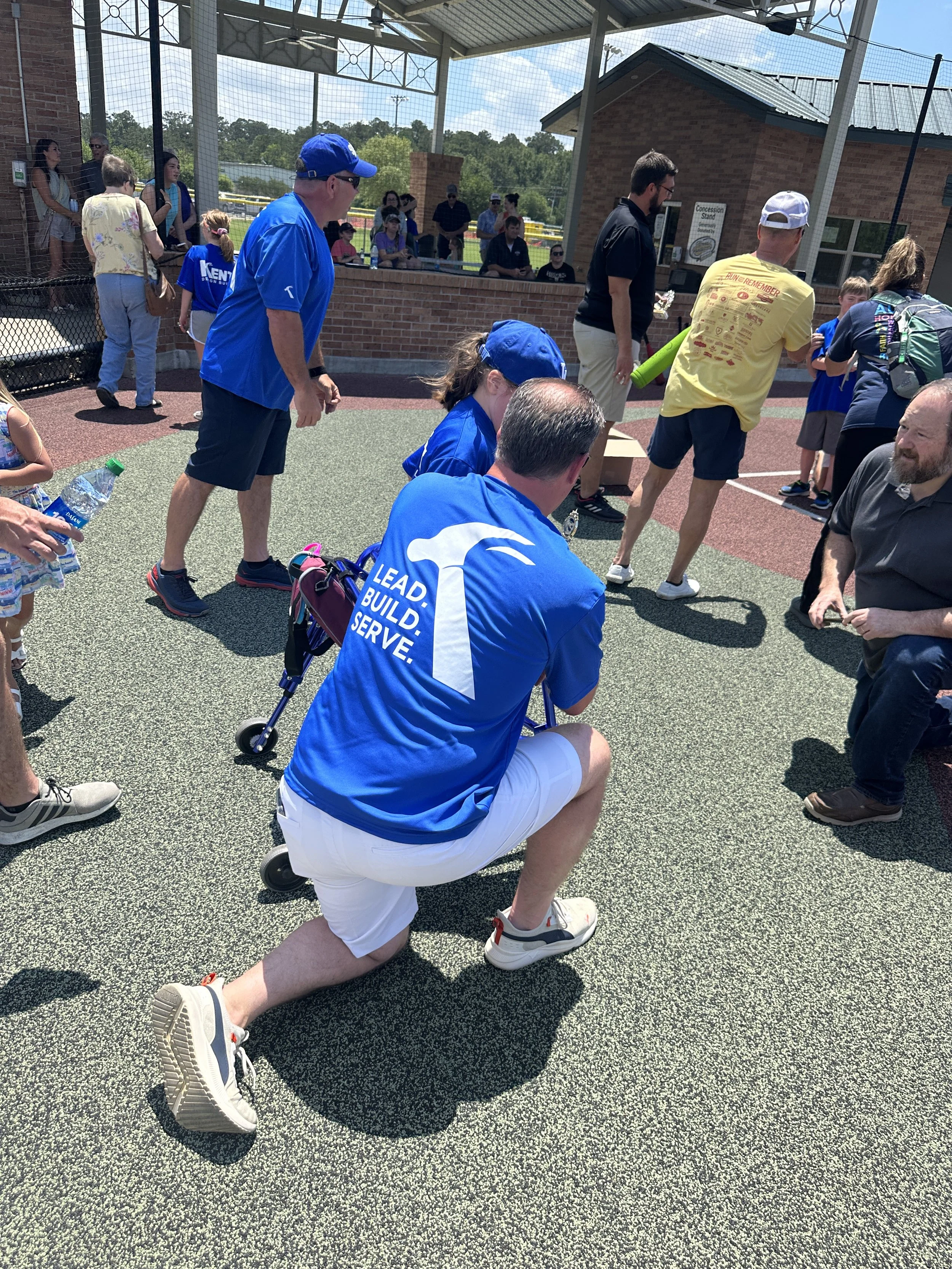 A group of people, including children and adults, gather outdoors on a sports court near a brick building with a concession stand, some are wearing blue jackets and shirts, and a man is kneeling on one knee in the foreground wearing a blue shirt with