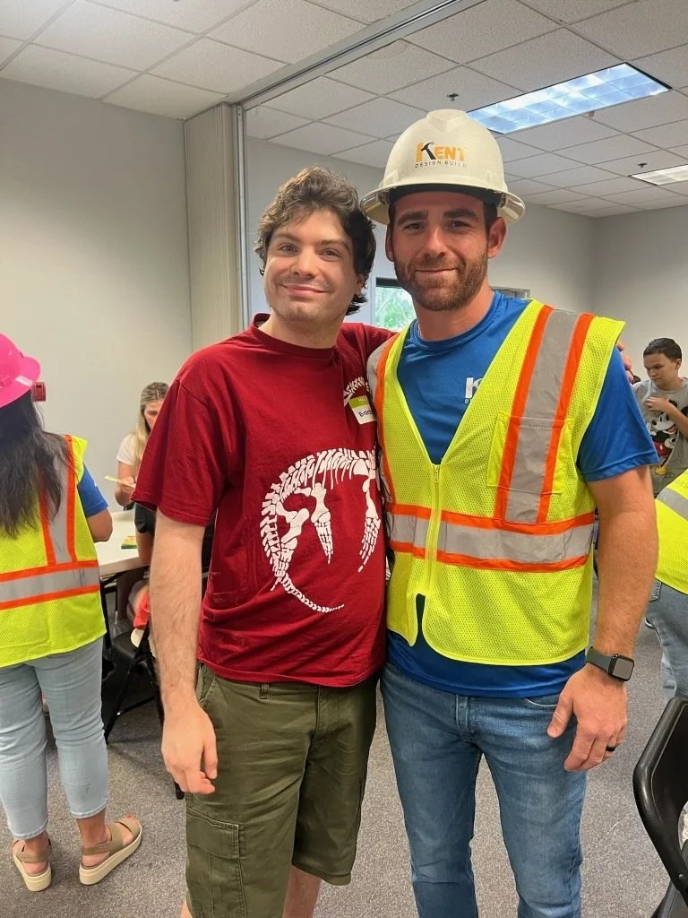 Two men standing together, smiling. One wearing a red T-shirt with a white dinosaur skeleton graphic, the other in a high visibility safety vest, blue T-shirt, and a white construction helmet with a logo. Several people in safety vests are in the bac