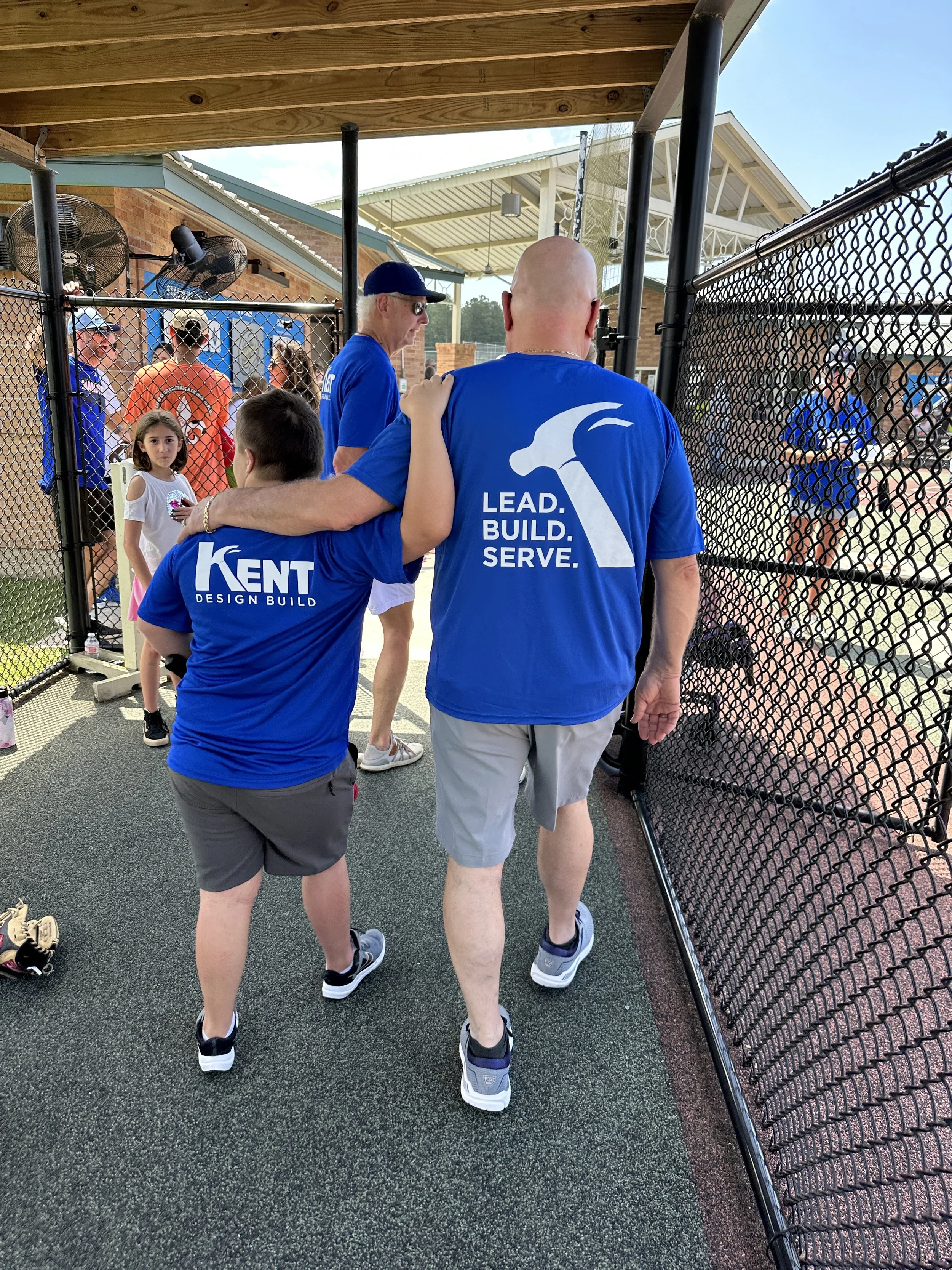 Two people in blue shirts with "Kent Design Build" and "Lead Build Serve" slogans, walking past a fence onto a sports field, with children and other adults in the background.