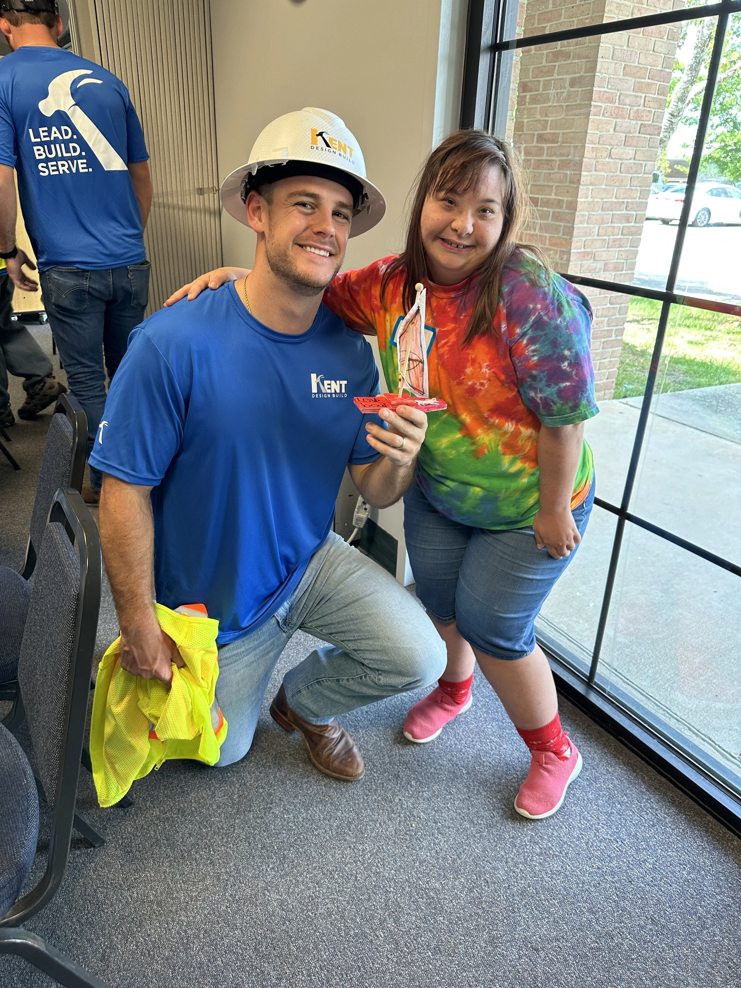 A man wearing a blue shirt and a safety helmet kneels next to a girl in a tie-dye shirt, both smiling and holding a small sailboat craft. The setting is indoors near large windows, with other people present.