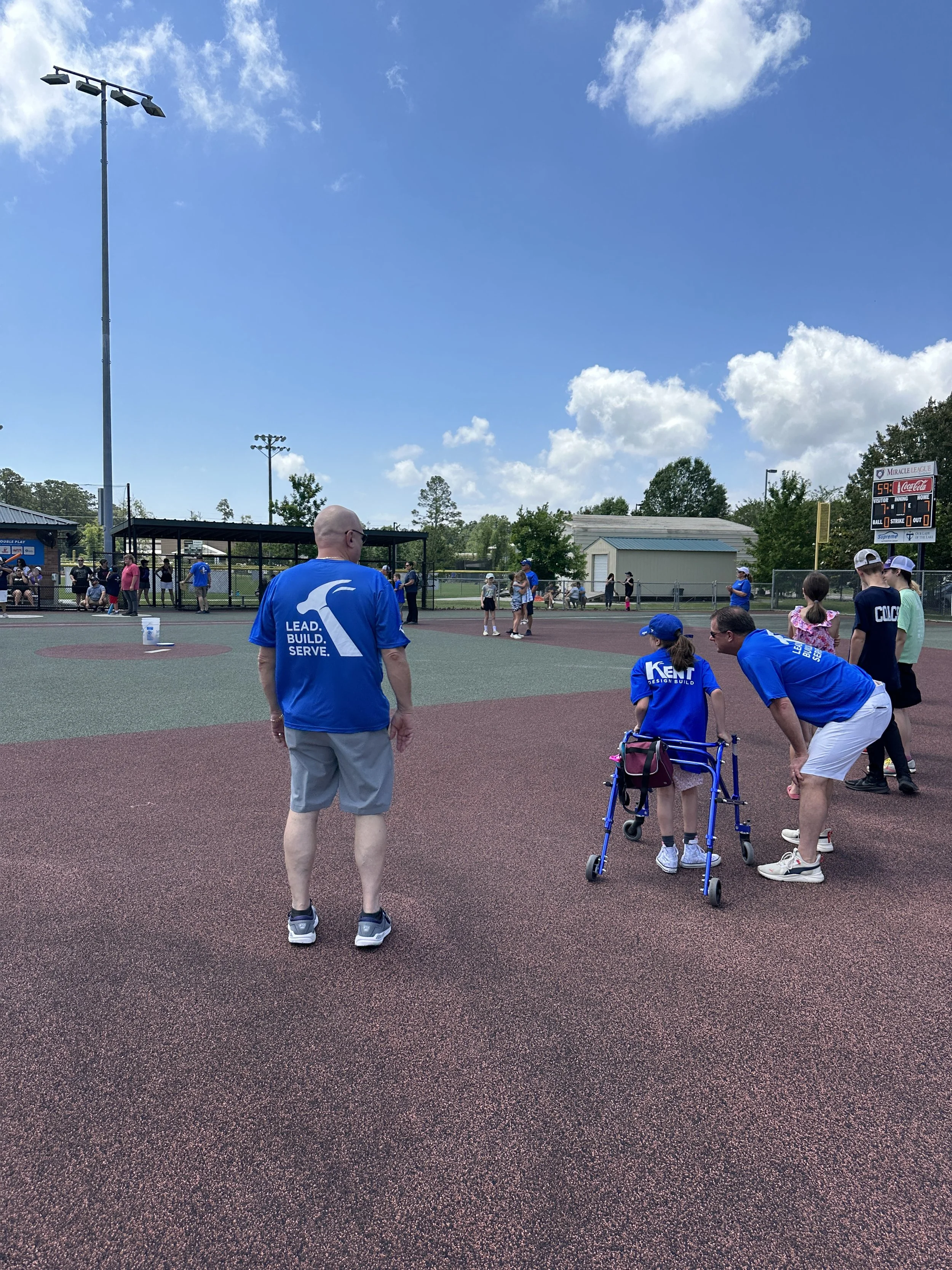 People at a baseball field, including a young girl using a walker, with a scoreboard and blue sky overhead.