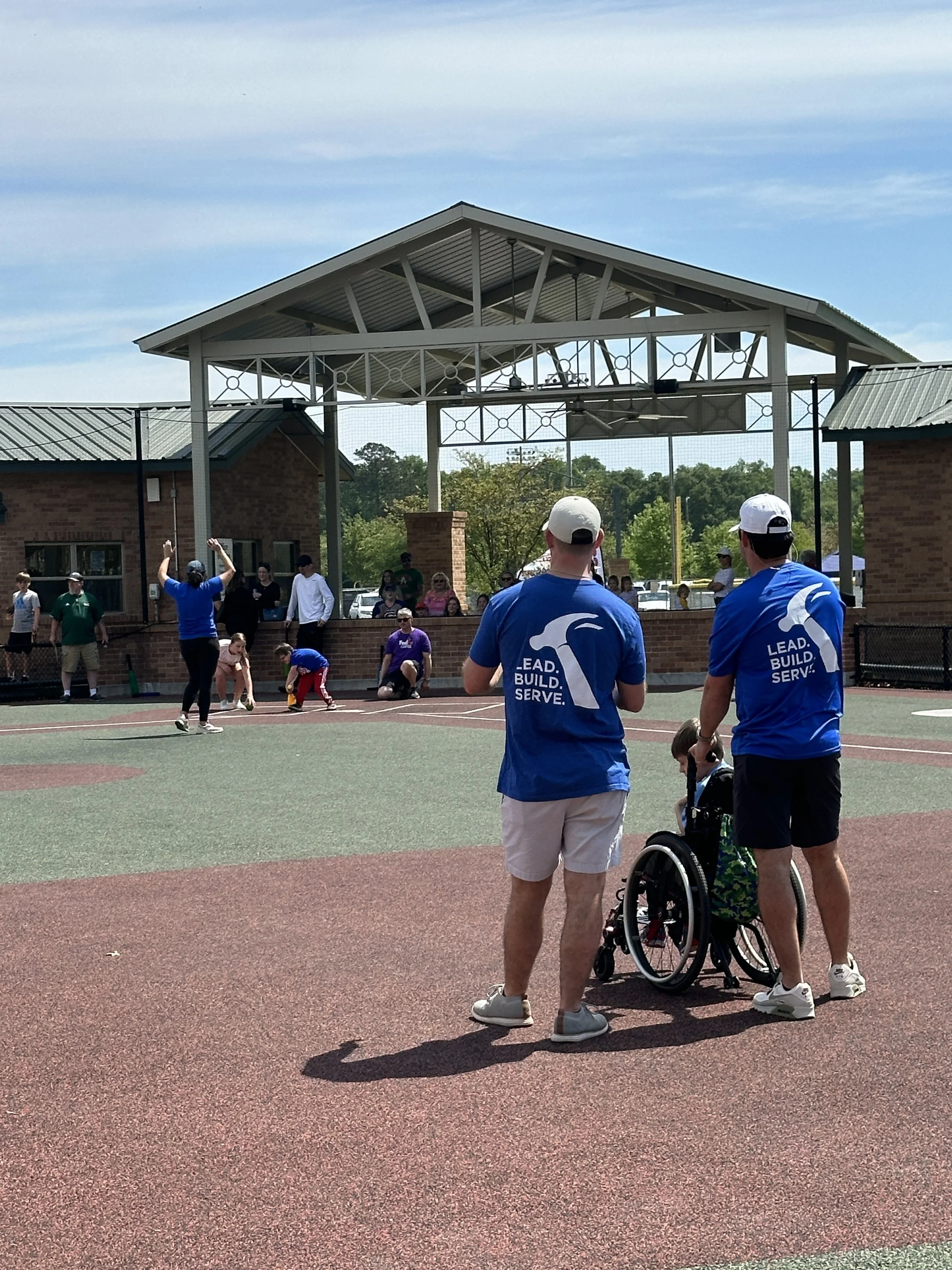 People playing basketball outdoors, with two volunteers from the YMCA observing, one in a wheelchair, in a park with a covered pavilion and trees in the background.