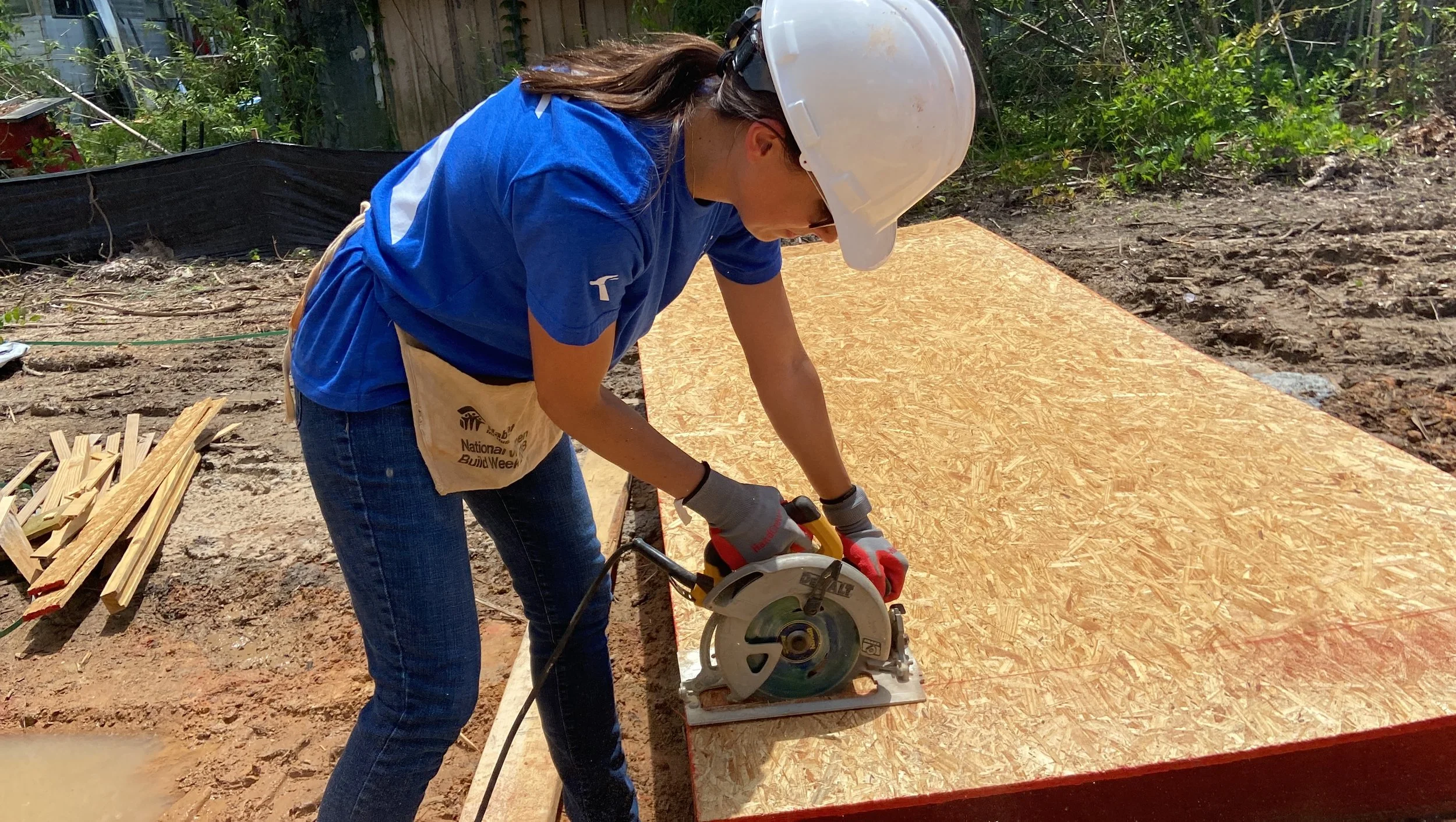 A woman wearing a white safety helmet, blue T-shirt, gray gloves, and jeans uses a circular saw to cut a large wooden panel outdoors. She is leaning over the panel on a construction site with greenery in the background.