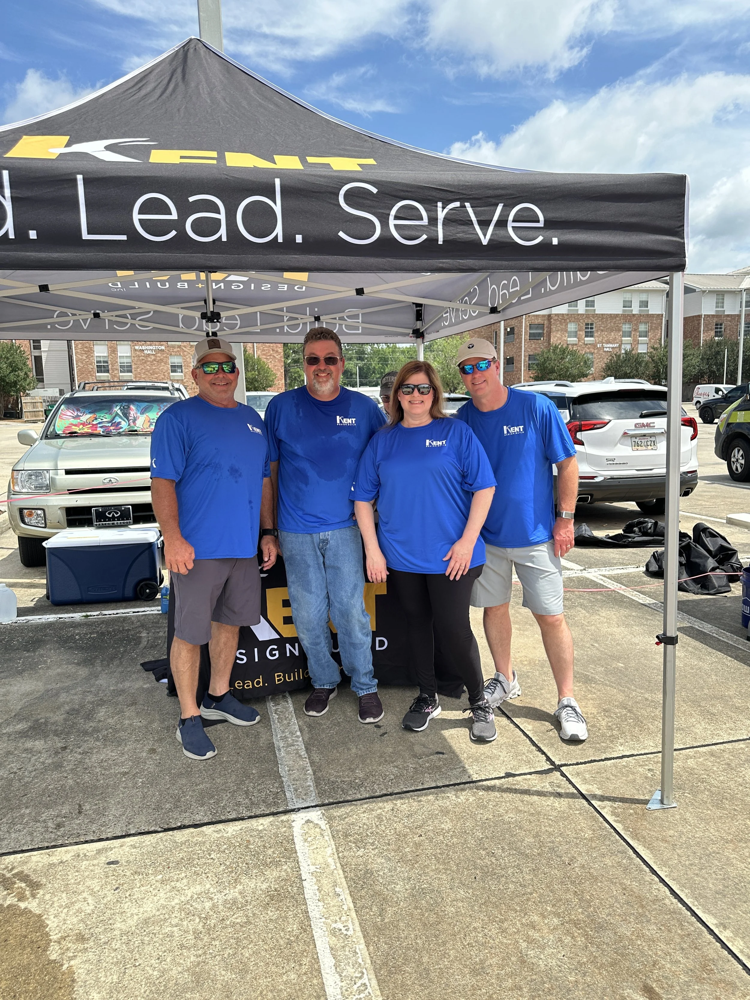 Four people standing under a black tent with 'Kent' branding, all wearing matching blue shirts. They are in a parking lot with cars in the background, smiling at the camera.