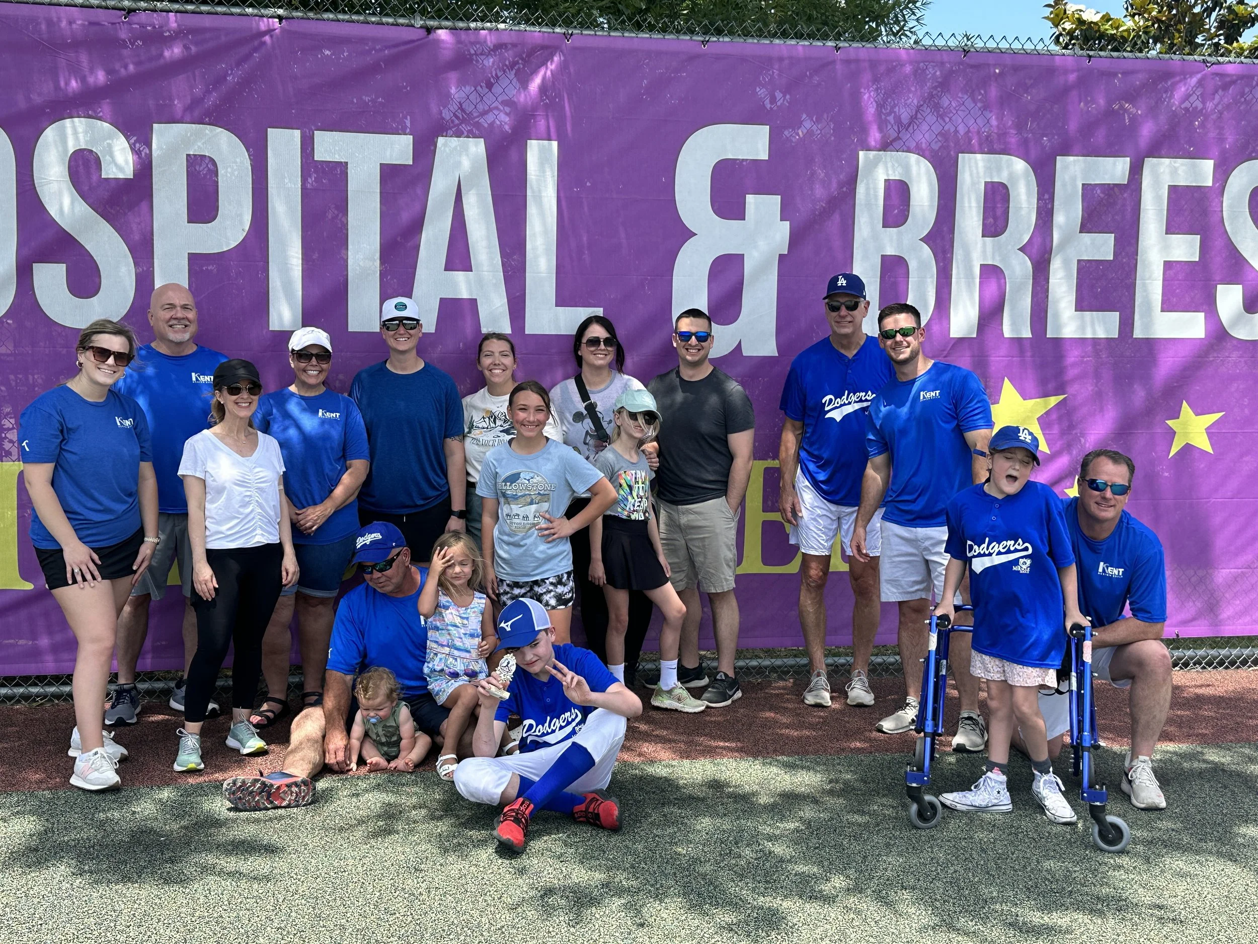 A group of people, including children and adults, poses for a photo in front of a large purple banner with white and yellow text that reads "Hospital & Breezes". Some individuals are wearing blue Dodgers shirts, and others are dressed casually. The g