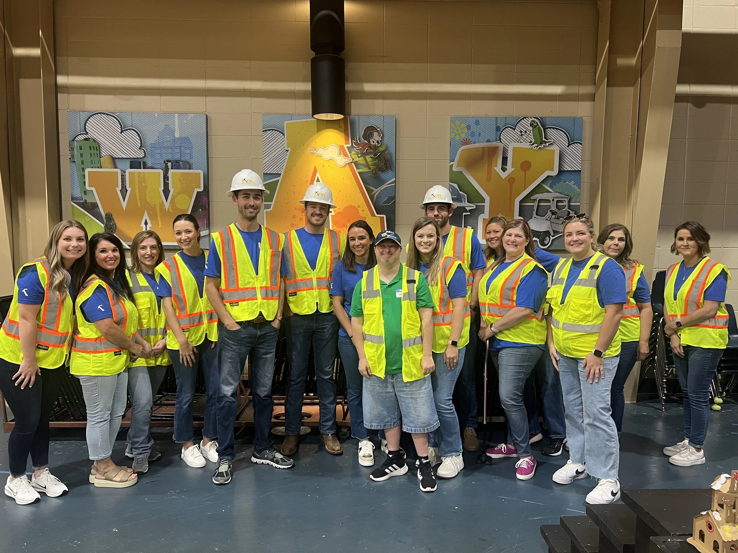 Group of people in yellow safety vests and helmets posing in front of large colorful art background with the words 'WAY' and 'W'.