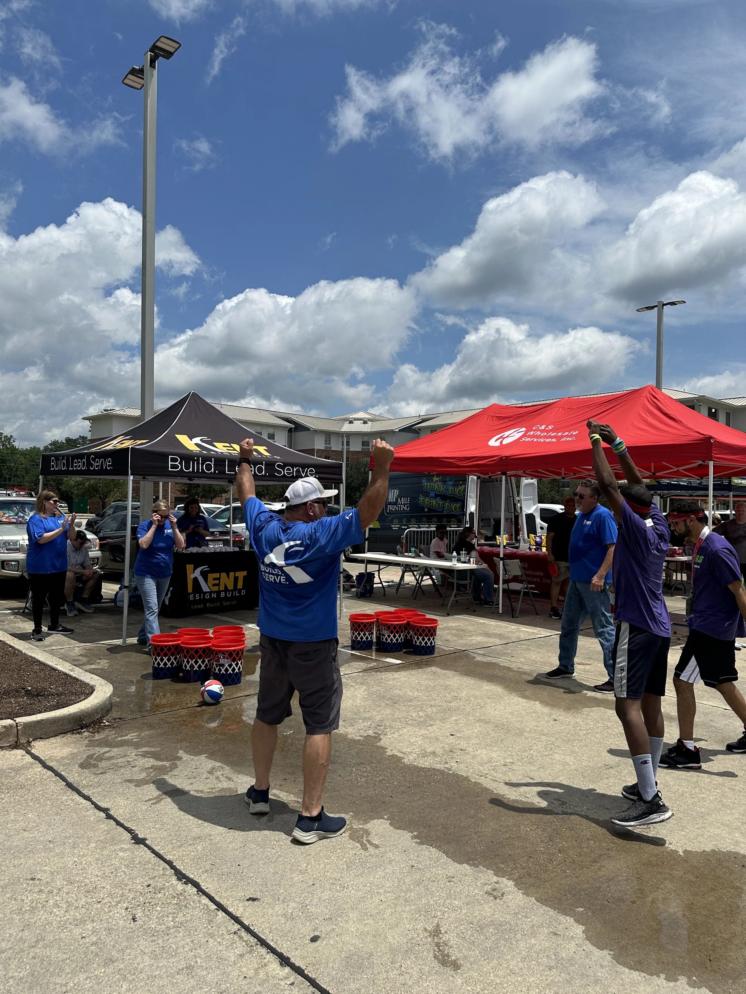 People playing a water balloon toss game outdoors under tents, with clouds and a blue sky overhead.
