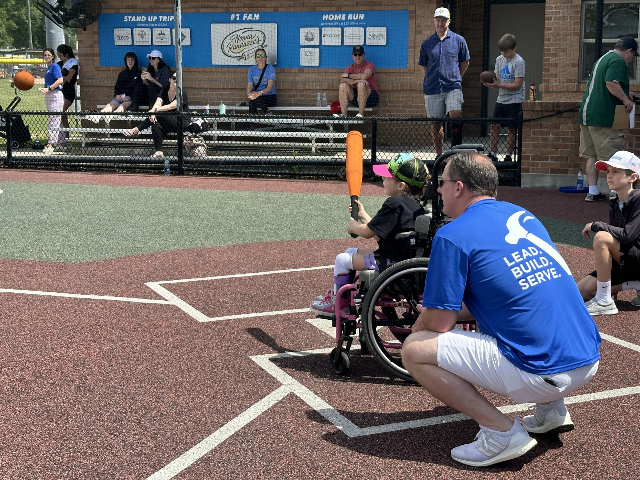Young girl in a wheelchair with a pink baseball cap, holding an orange baseball bat, next to an adult man in a blue shirt and white shorts, participating in a baseball game at a sports field. Spectators sit behind a fence in the background.