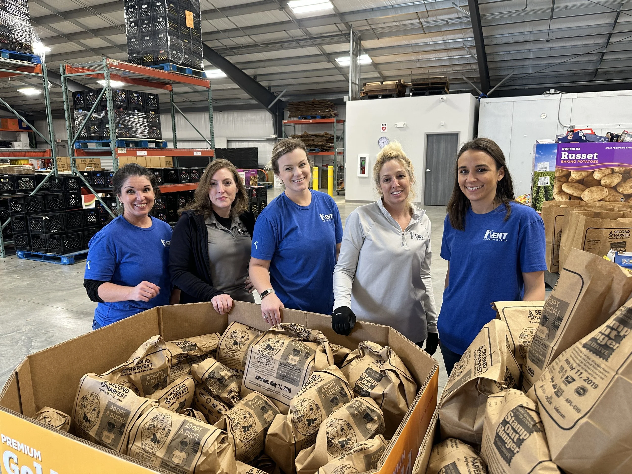 Five women standing in a warehouse with shelves of crates and supplies in the background, smiling behind a large box of paper bags with printed text.