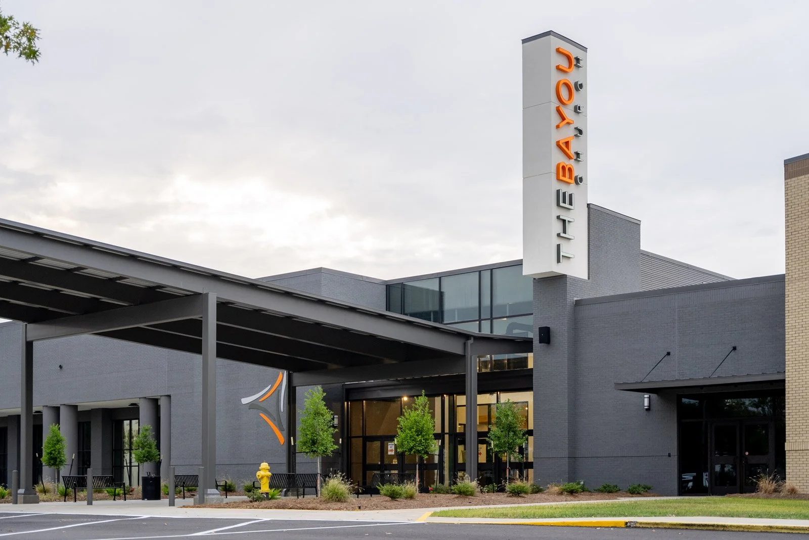 Exterior of The Bay restaurant with modern architecture, a tall vertical sign displaying the name, and a sidewalk with benches, trees, and yellow fire hydrant.