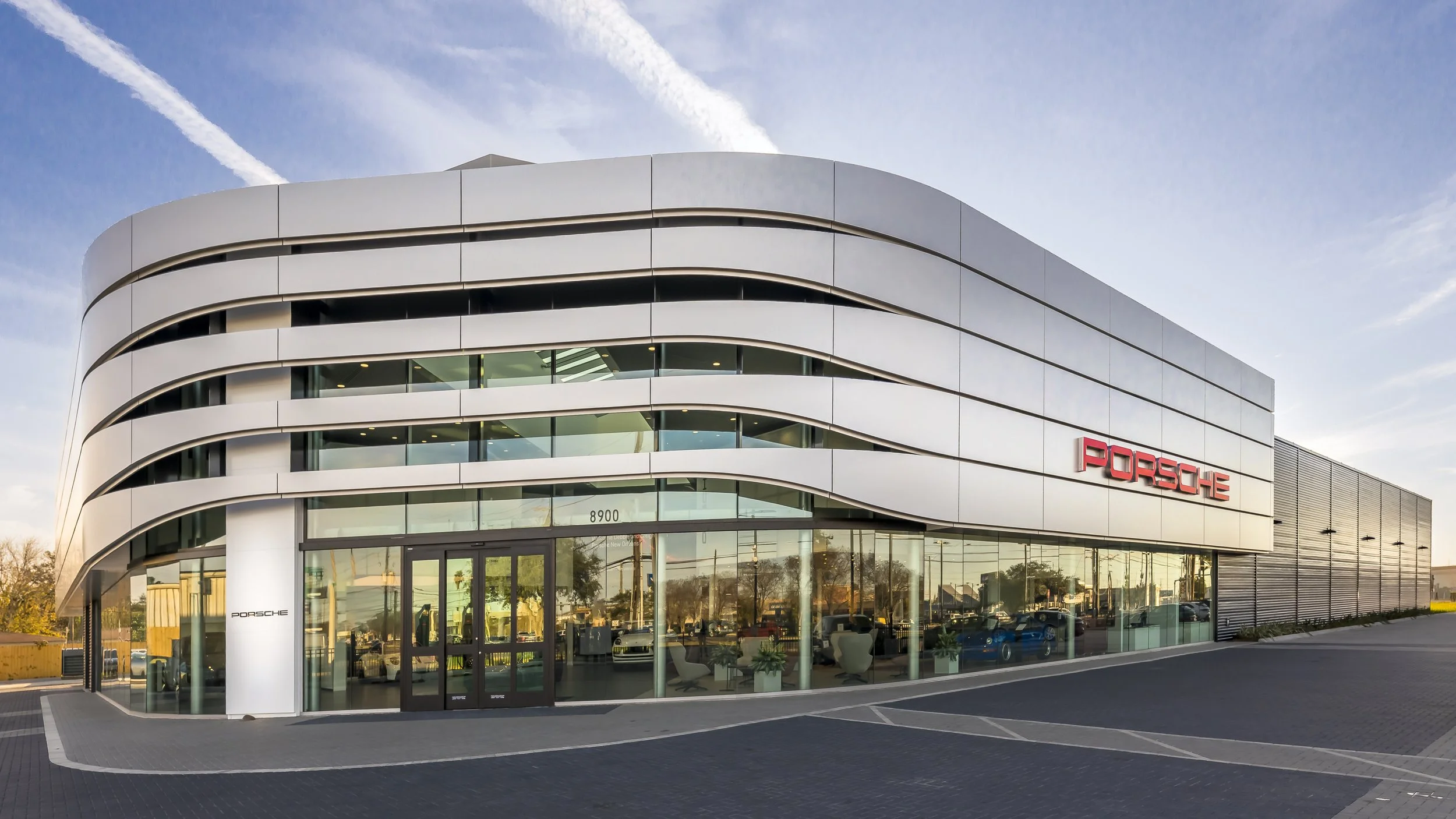 Modern Porsche dealership building with curved white exterior and large glass windows, featuring the Porsche logo, parked cars inside and outside, and a clear sky above.