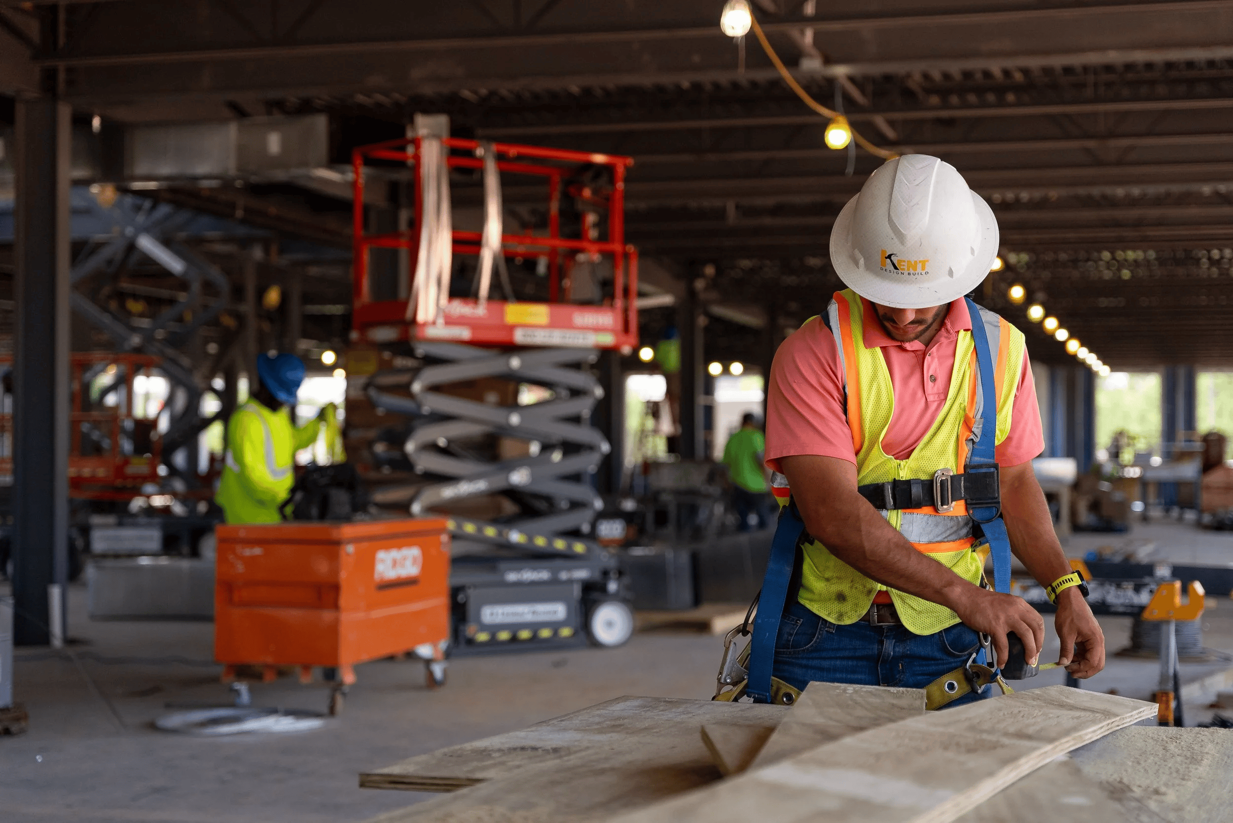 Construction worker wearing a safety helmet and vest working on a building project indoors with other workers and construction equipment in the background.