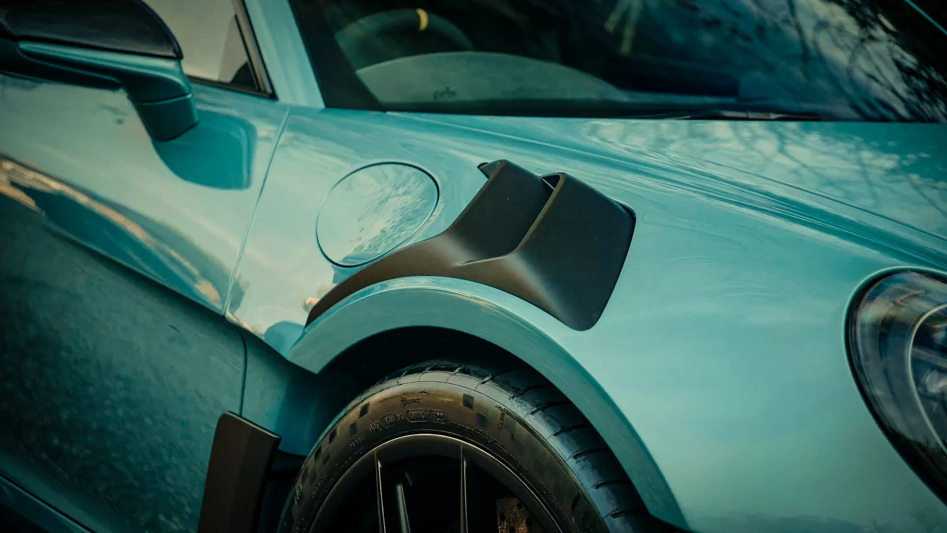 Close-up of a light blue vintage car's front side, showing part of the windshield, side mirror, tire, and headlight with reflections of trees and sky.