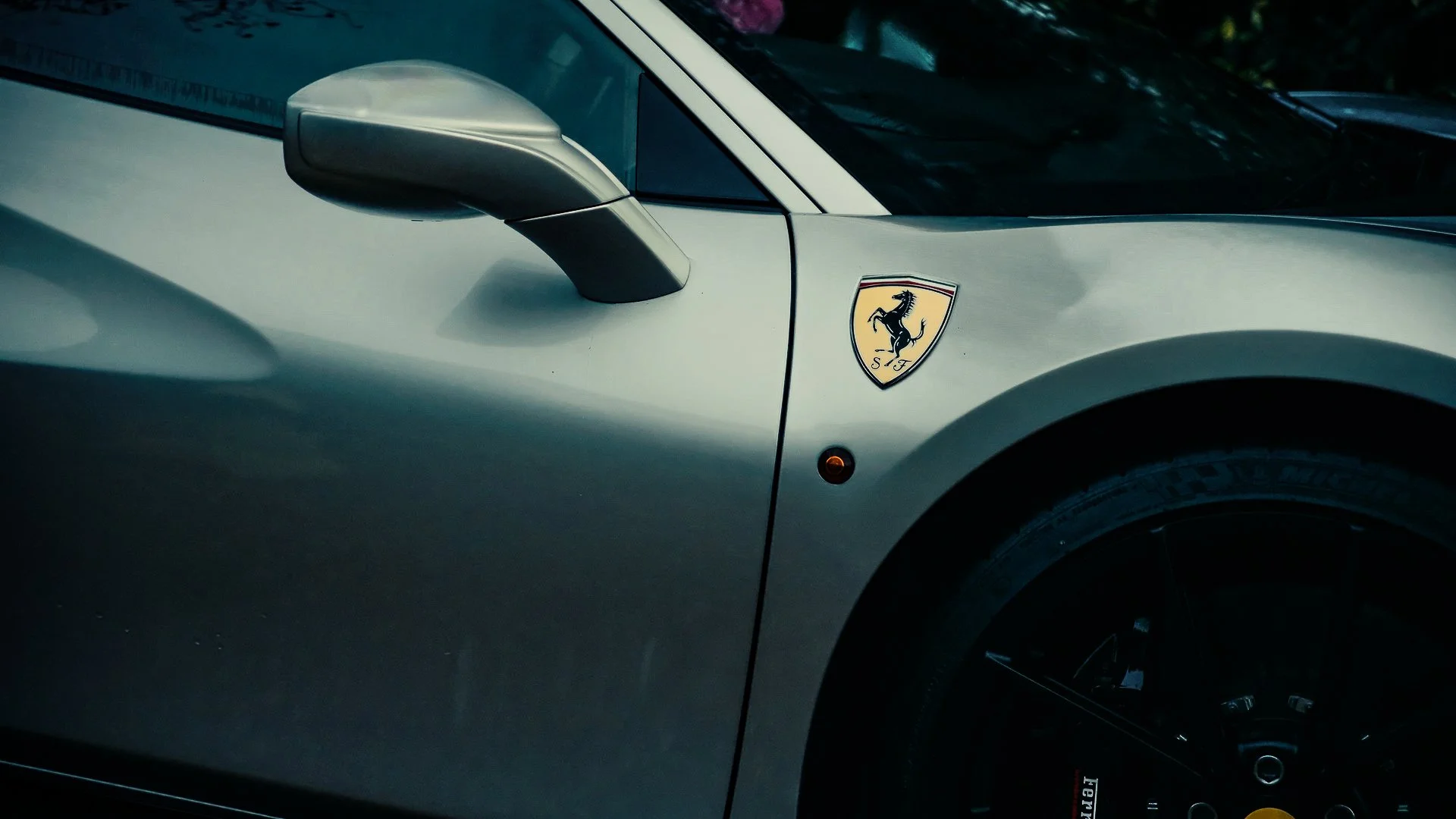 Close-up of a silver Ferrari sports car, showing part of the front end with a side mirror, Ferrari badge, and wheel.