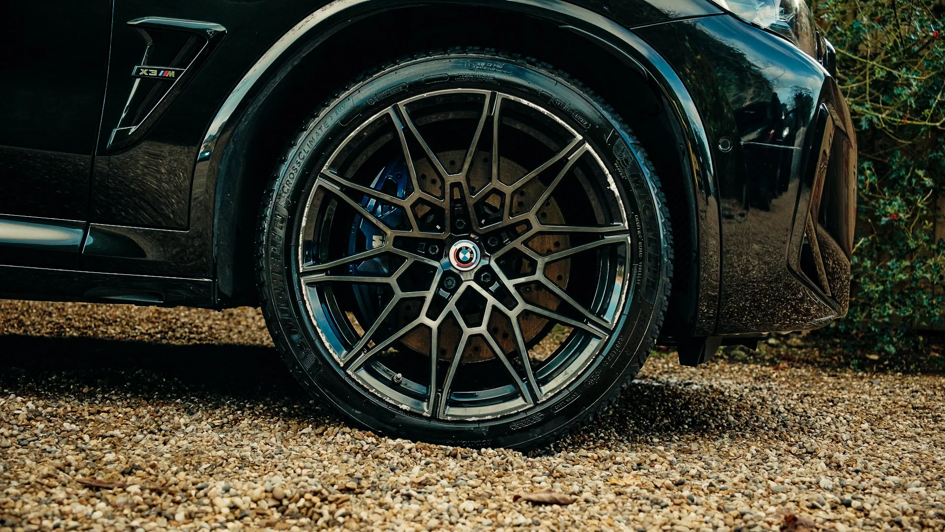 Close-up of a black BMW car's wheel with a detailed, multi-spoke rim and a BMW logo in the center, parked on gravel with some greenery in the background.