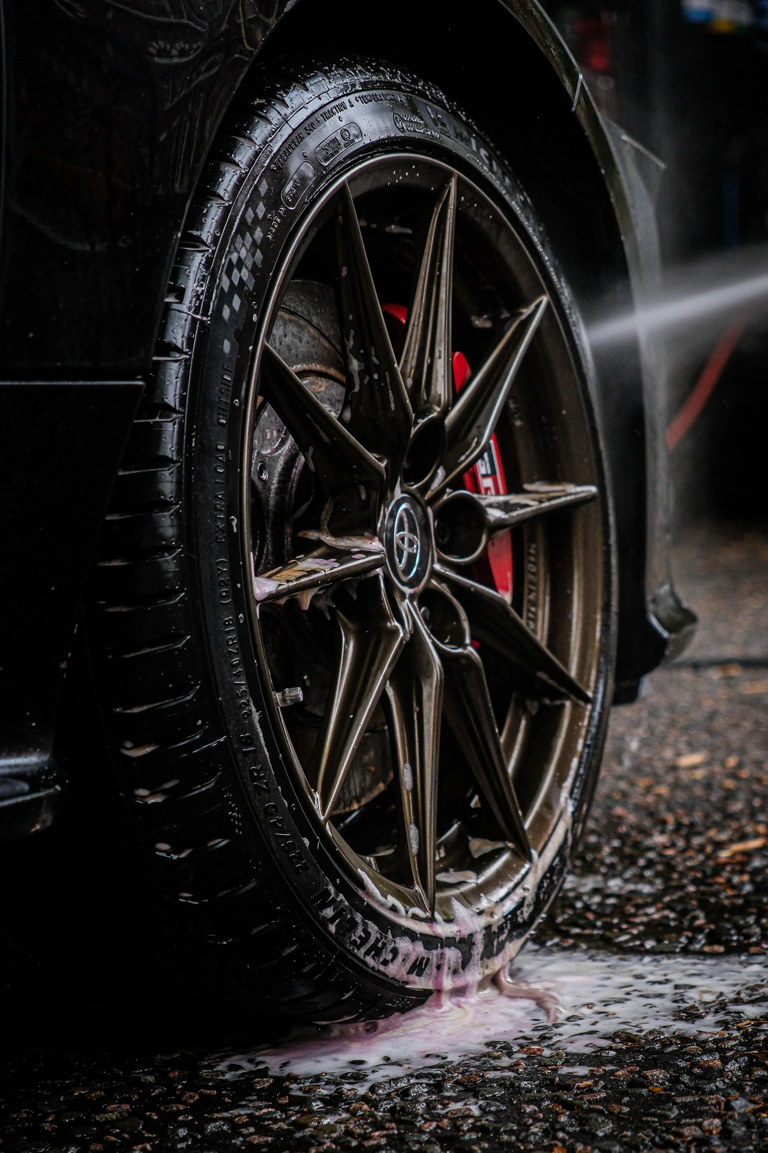 Close-up of a black car tire being washed with soap and water, with a red brake caliper visible behind the alloy wheel.