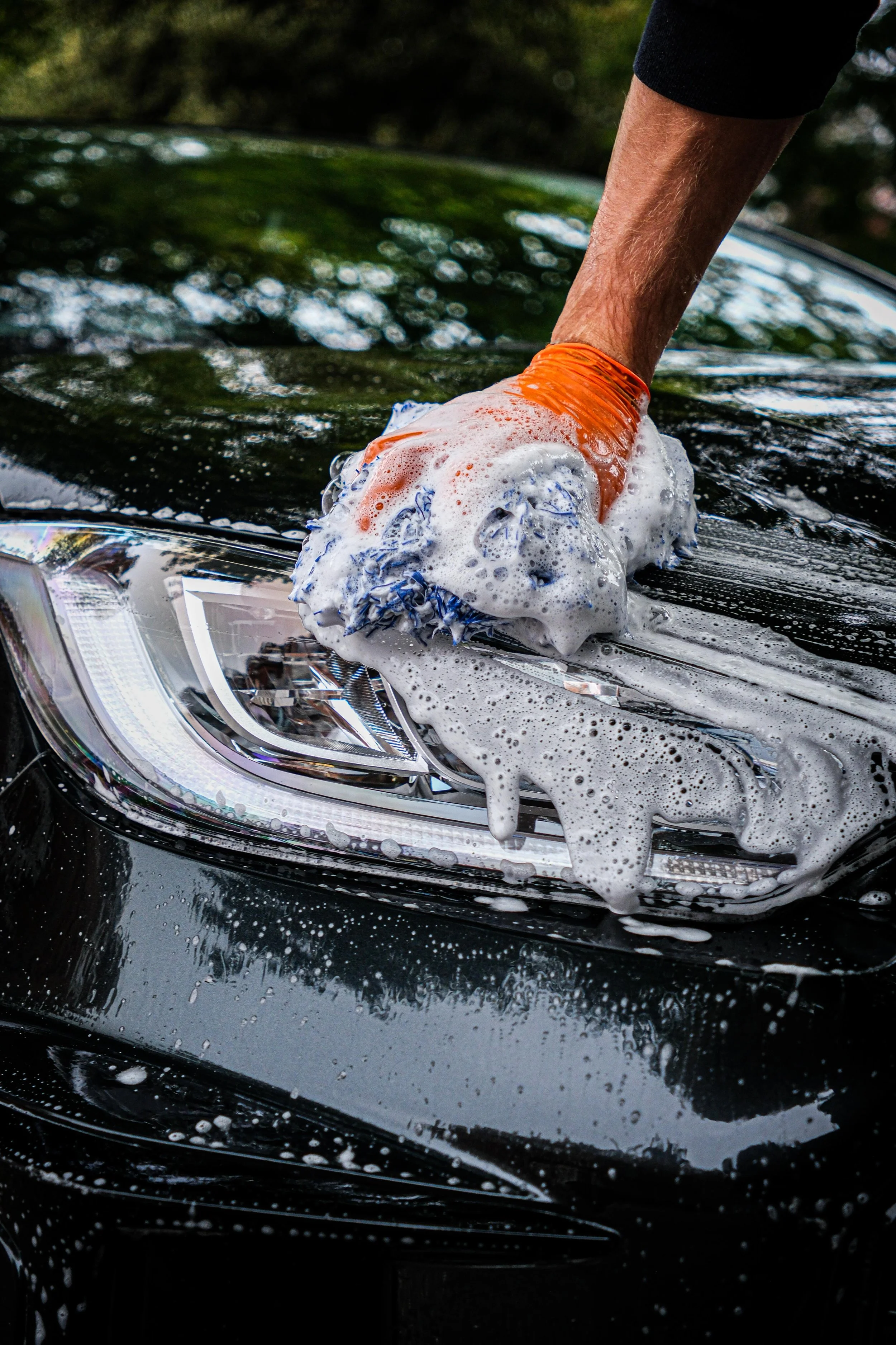 Person washing a black car with soap and sponge outdoors, wearing an orange glove.