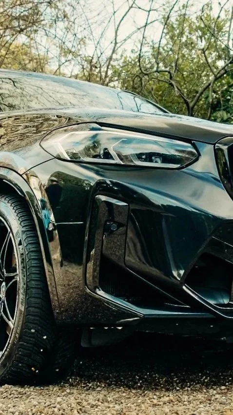 Close-up of the front of a black sports car parked outdoors with trees and sky in the background.