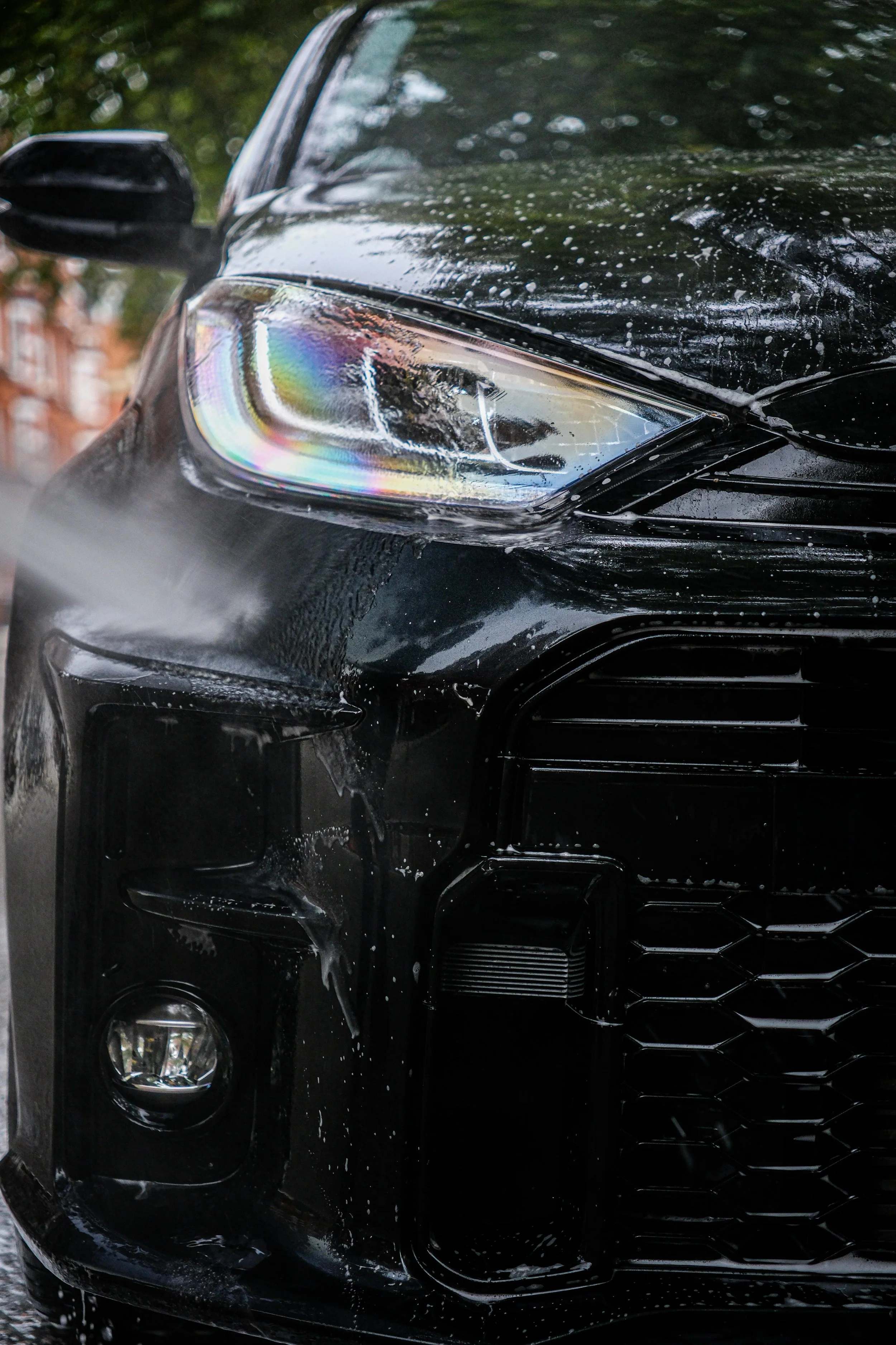 Close-up of the front of a black car with water and soap on the surface, showing the headlight, grille, and side mirror.