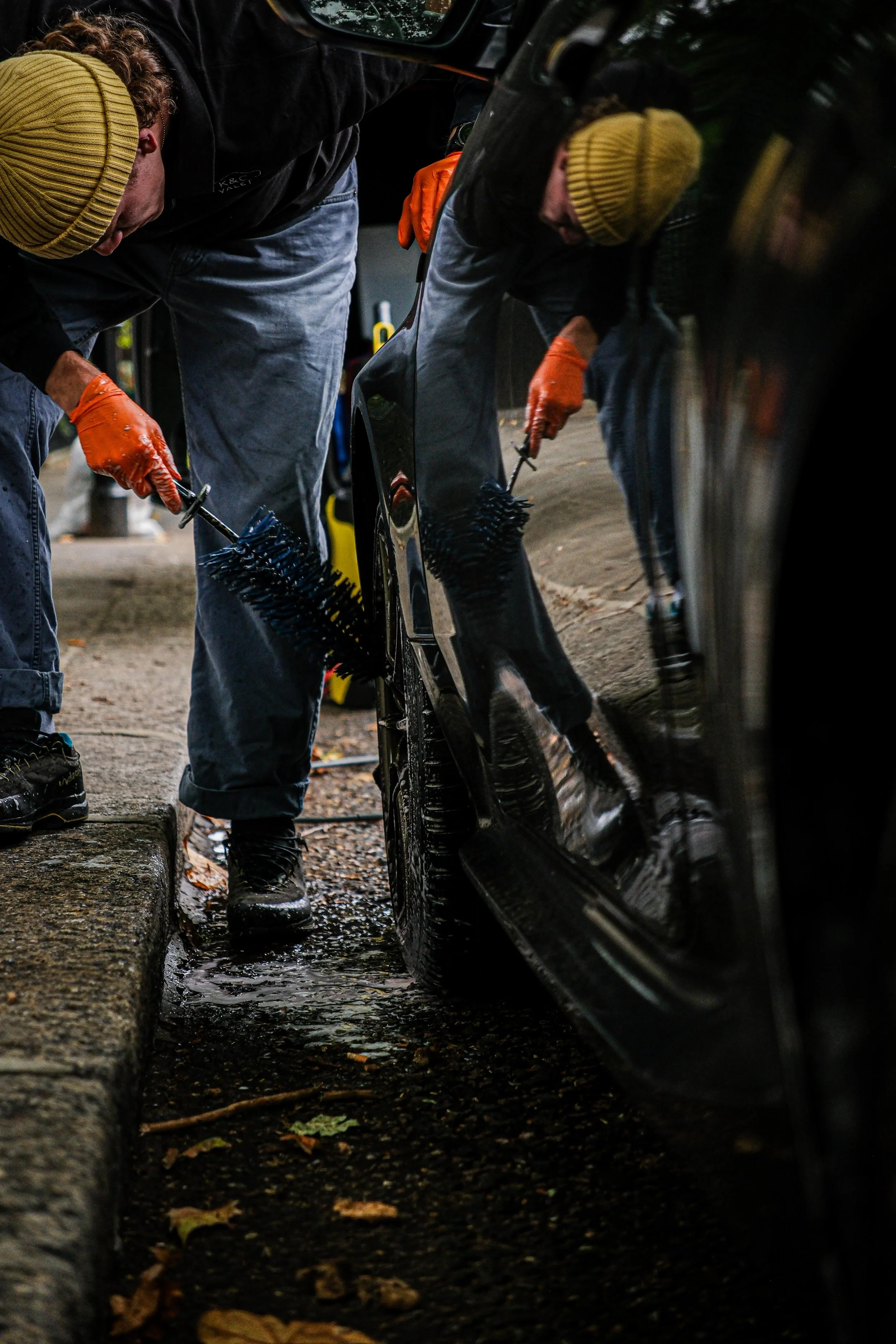 Two people cleaning the side of a black car with brushes, wearing gloves and yellow beanies.